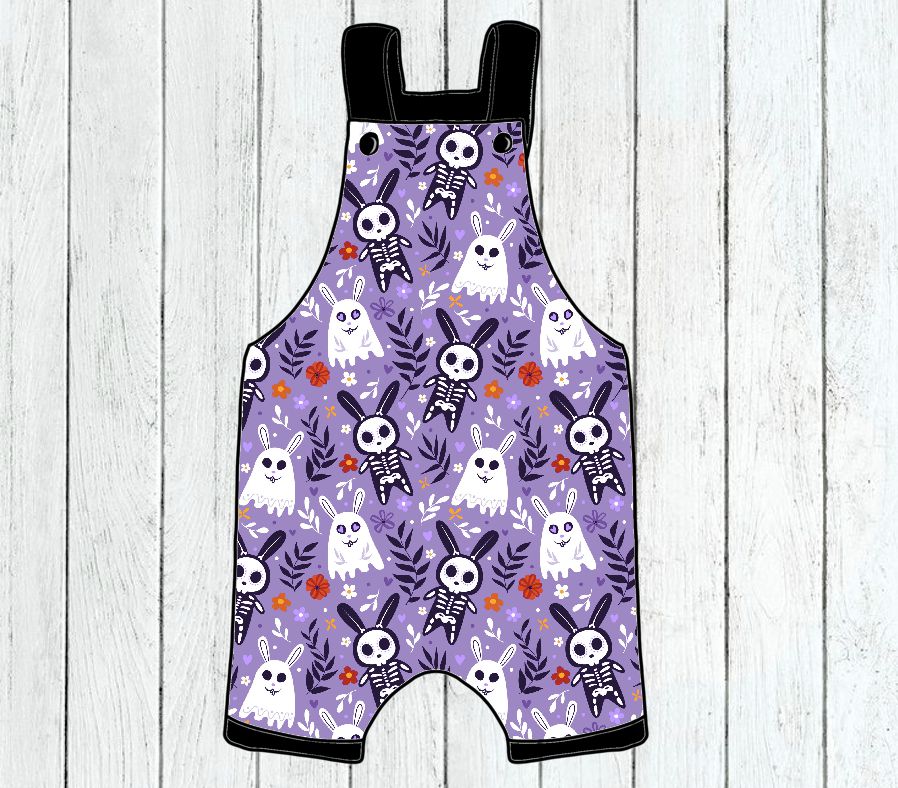 Macacão infantil roxo com padrão de Halloween de fantasmas e esqueletos em fundo de madeira branca.