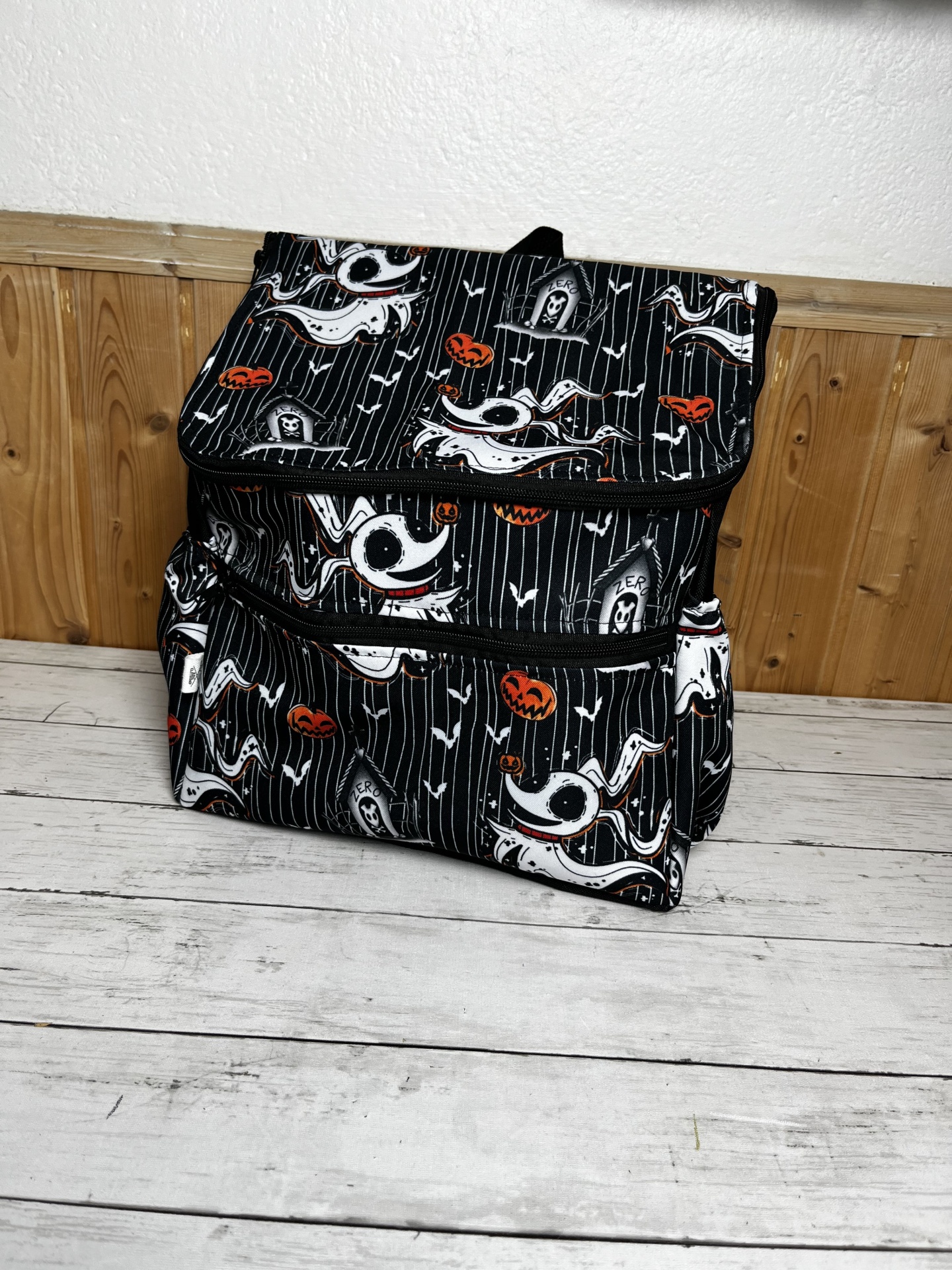 Mochila preta com padrão de fantasmas, abóboras e túmulos em ambiente interior com fundo de madeira e parede branca