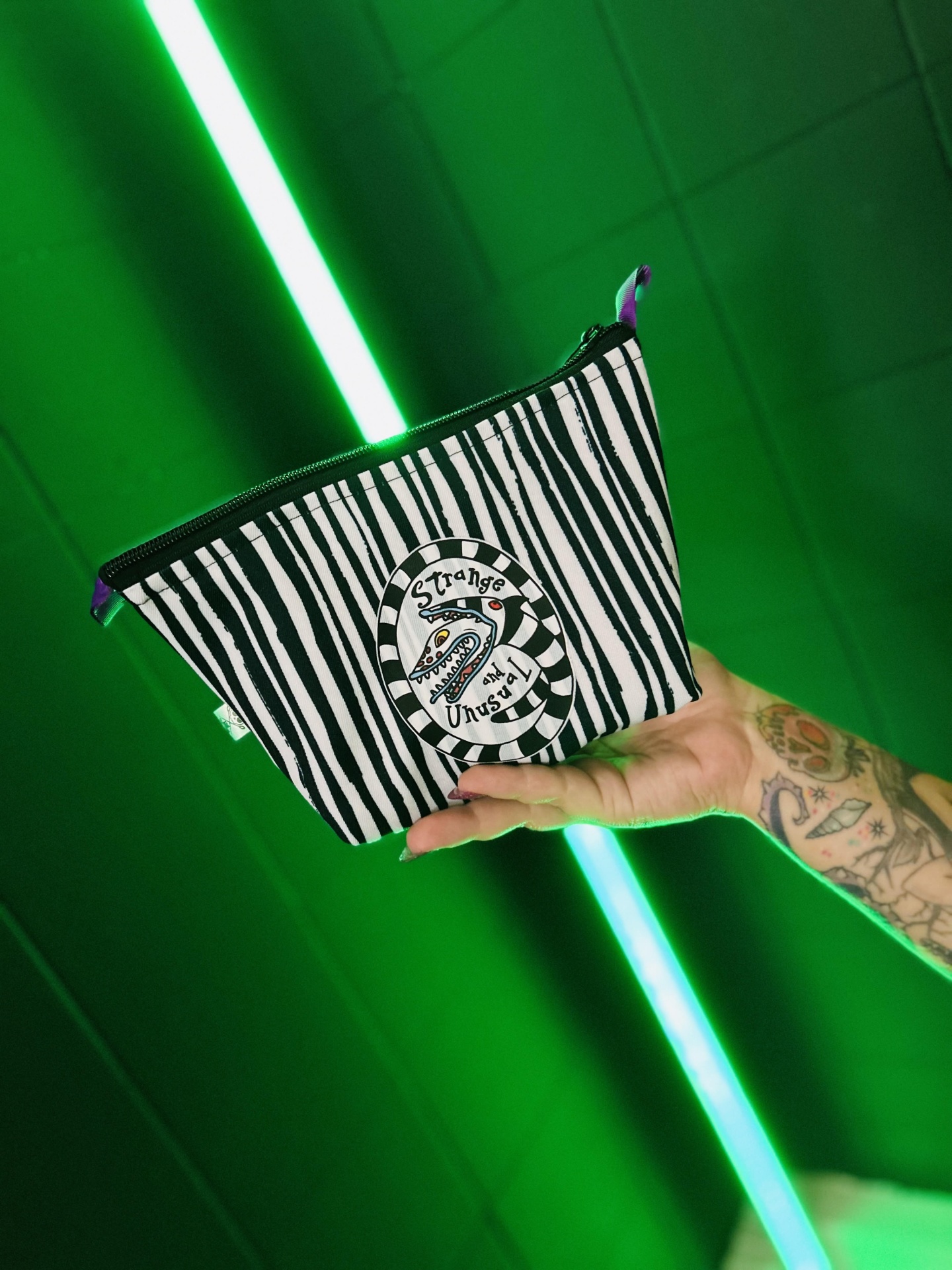 Bolsa pequena preta e branca com emblema 'Strange Unusual', mão tatuada a segurá-la sobre fundo verde com luzes