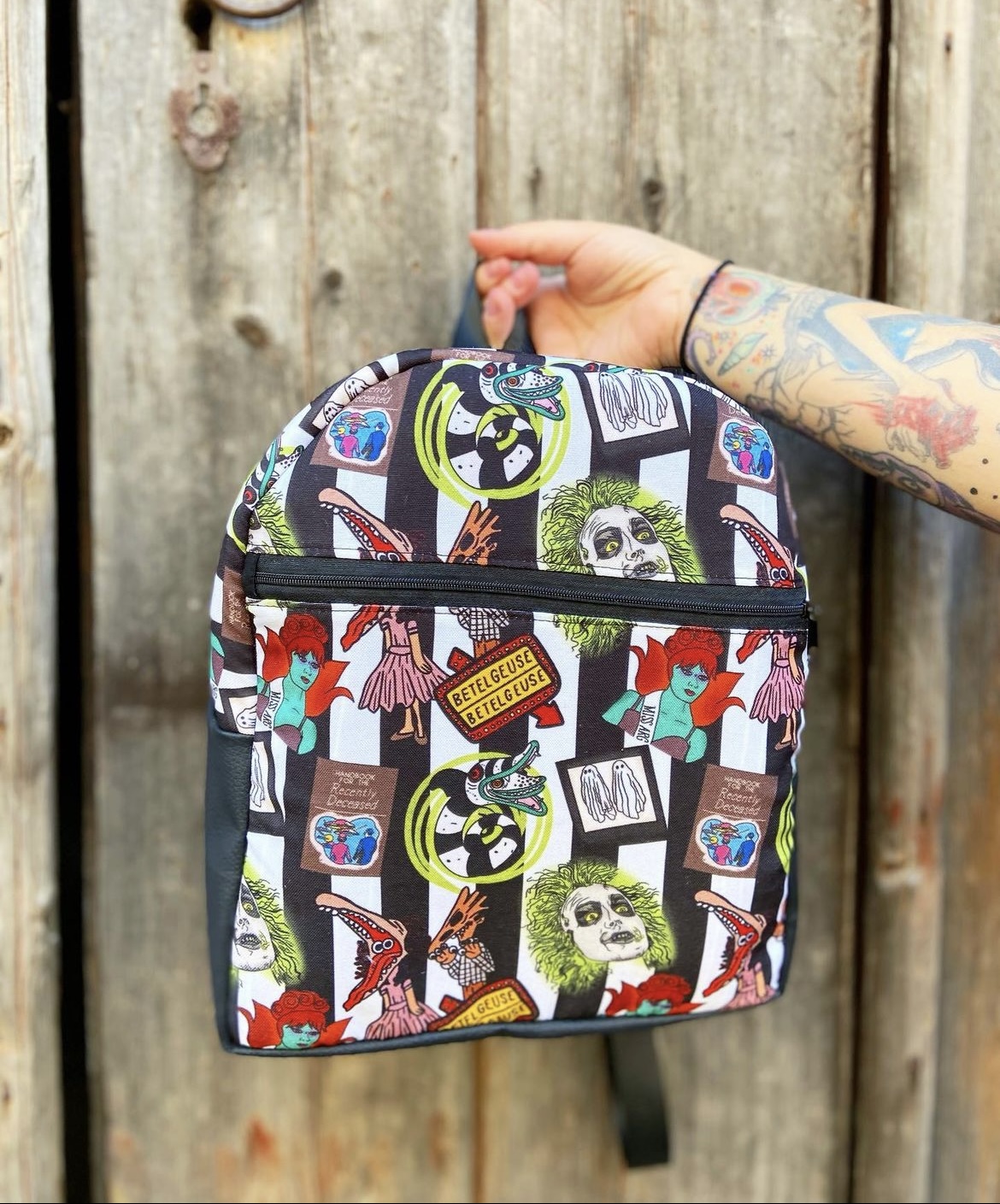 Mochila colorida com padrão de personagens e logótipos do filme Beetlejuice segurada por mão tatuada