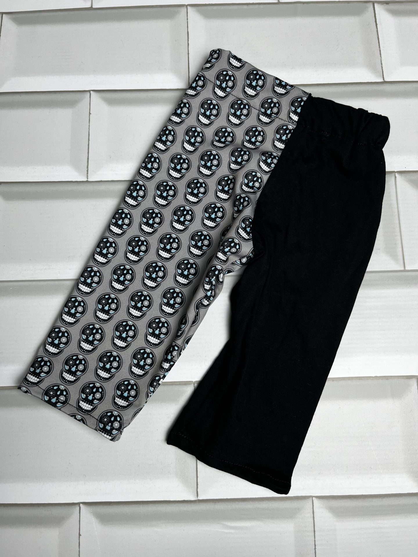 Calças infantis com perna preta e perna com padrão de caveiras sobre fundo branco de azulejos