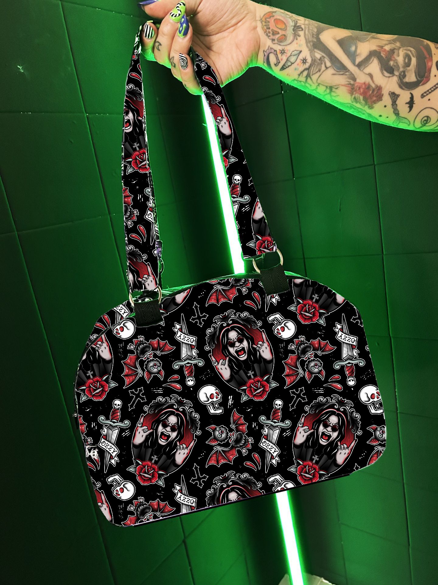 Mala de mão preta com padrão de caveiras, rosas e rostos femininos com uma mão tatuada segurando-a