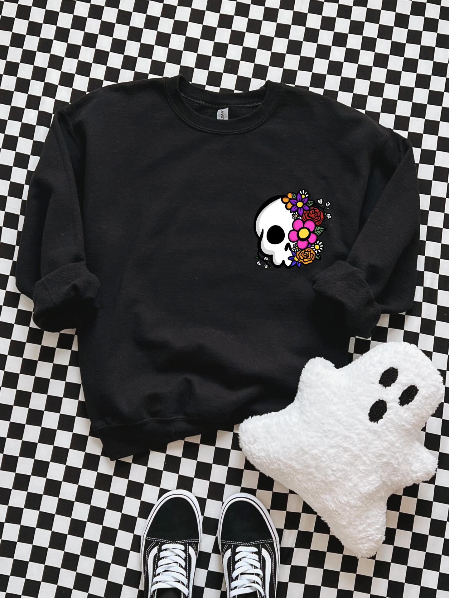 Sweatshirt preta com estampa de caveira flores coloridas, peluche fantasma branco, sapatilhas pretas sobre padrão quadriculado.