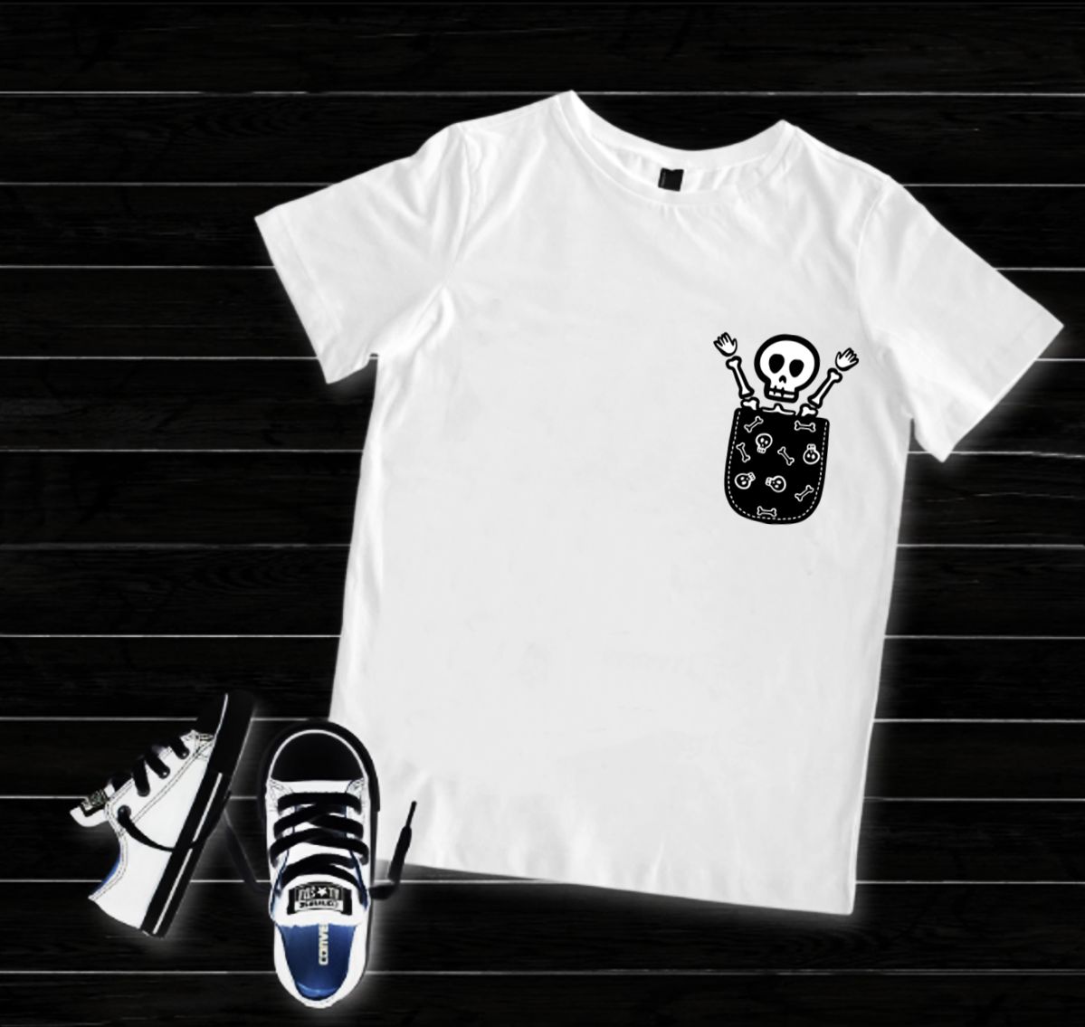 T-shirt branca com estampa de esqueleto no bolso, sapatilhas brancas e pretas ao lado sobre chão de madeira escura