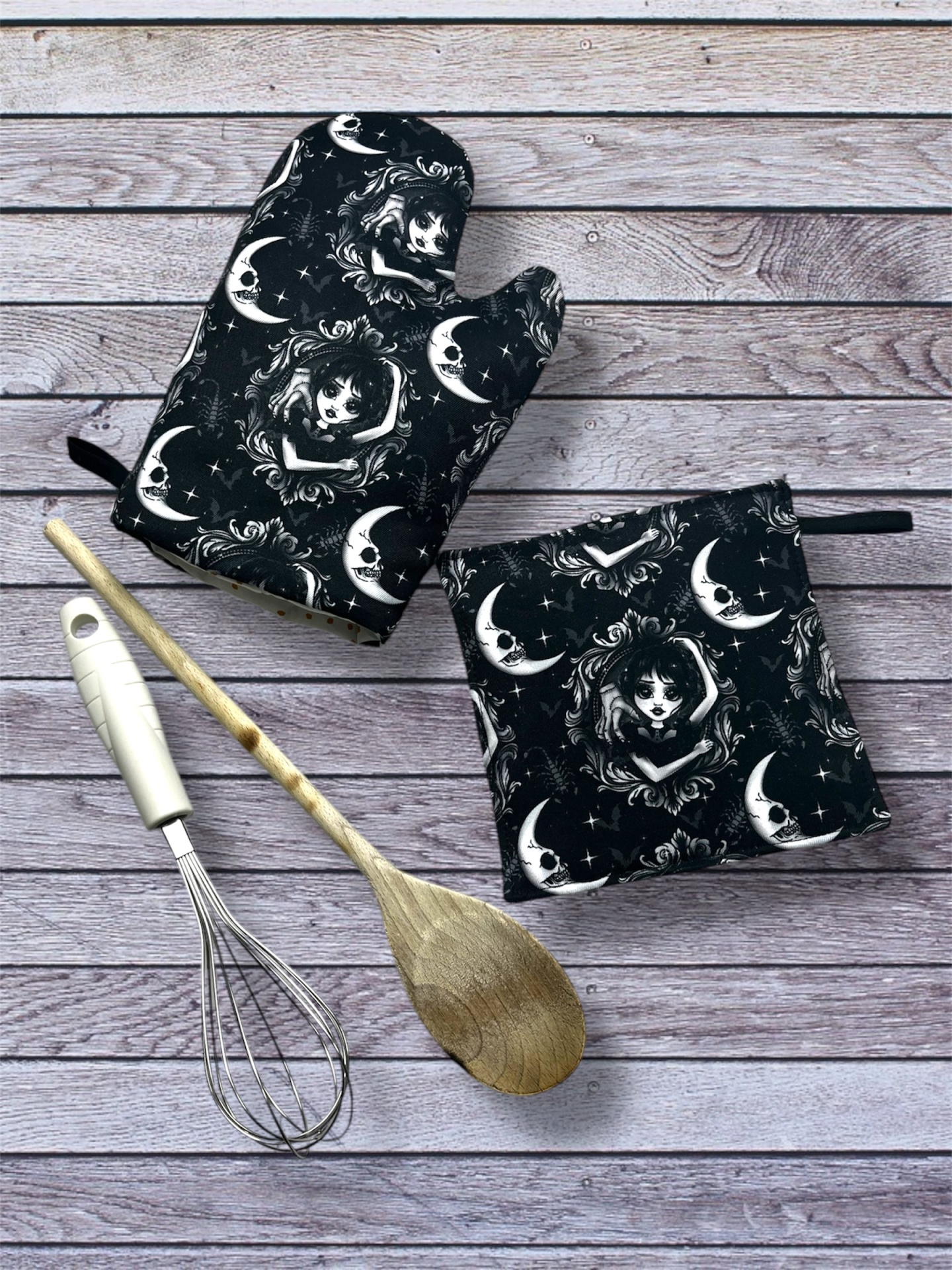 Conjunto de luva de forno e pega pretos com padrão lunar e rosto feminino ao lado de utensílios de cozinha sobre madeira cinzenta