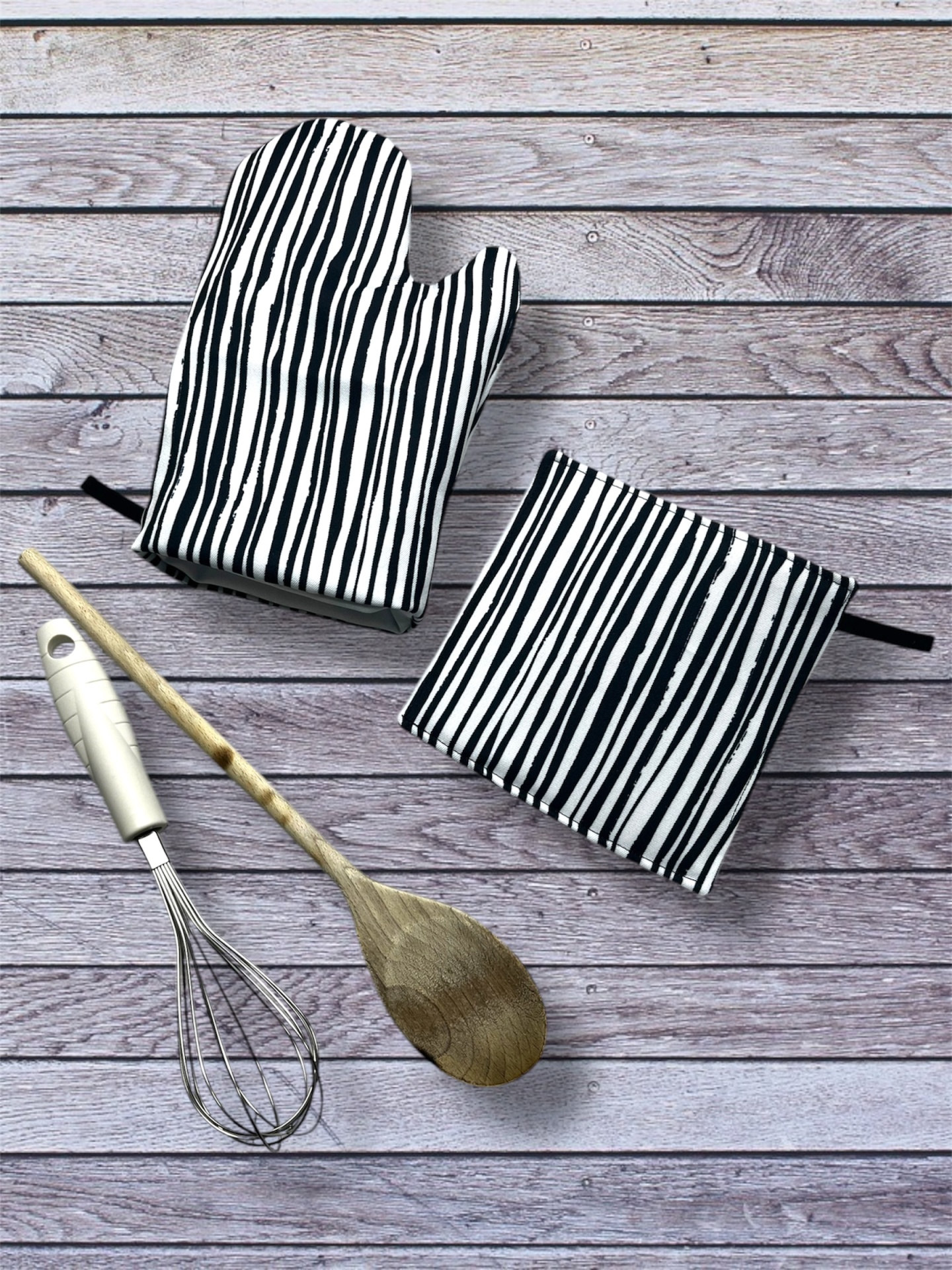 Luvas e descansos de panela com riscas preto e branco, batedor de metal e colher de pau sobre fundo de madeira
