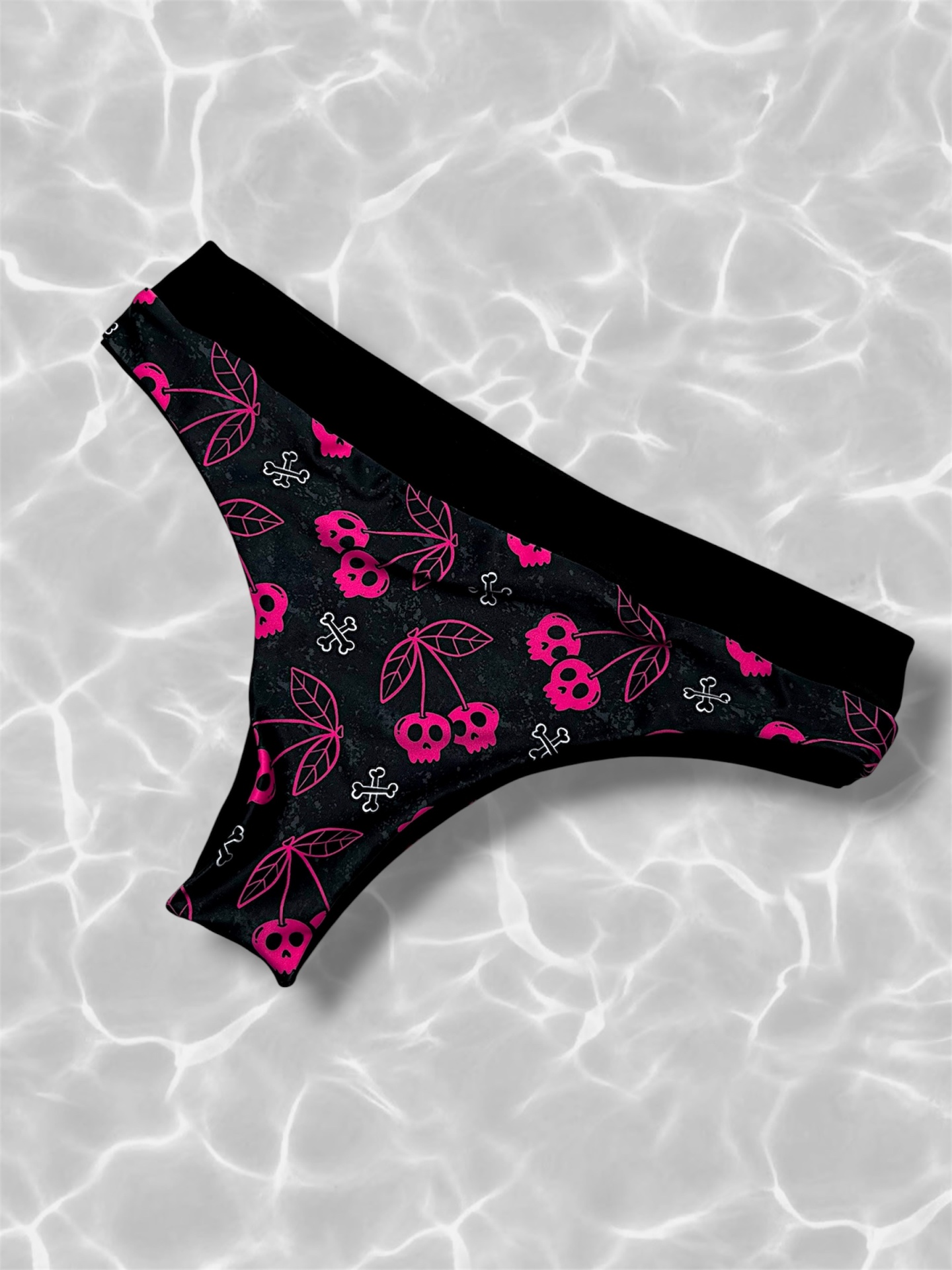 Parte de baixo de biquíni preto com estampado rosa de cerejas em forma de caveira e ossos, sobre fundo claro com reflexos de água.