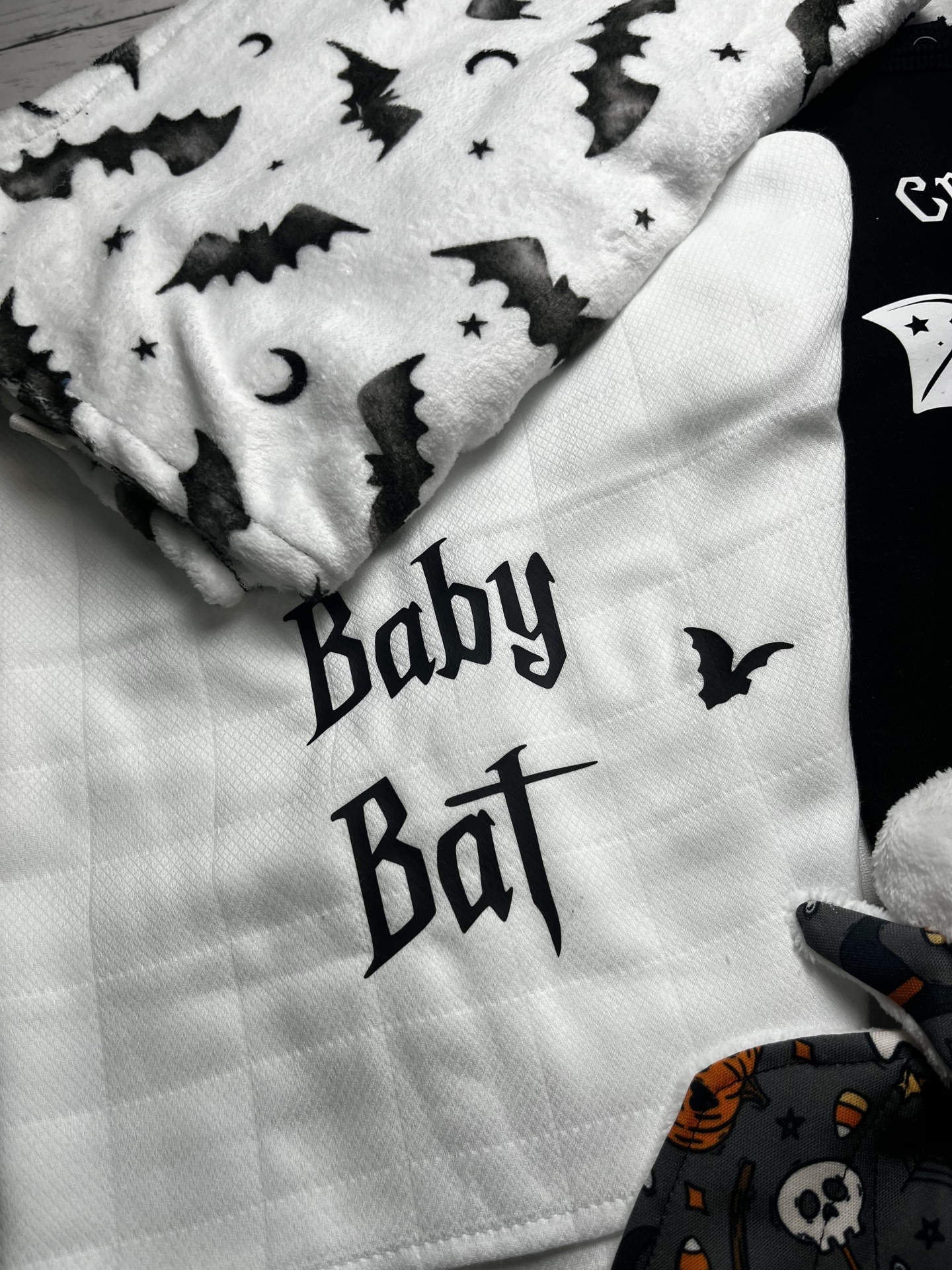 Pano branco com texto Baby Bat e padrão de morcegos preto e branco, ao lado de tecido com padrão Halloween em preto e laranja.