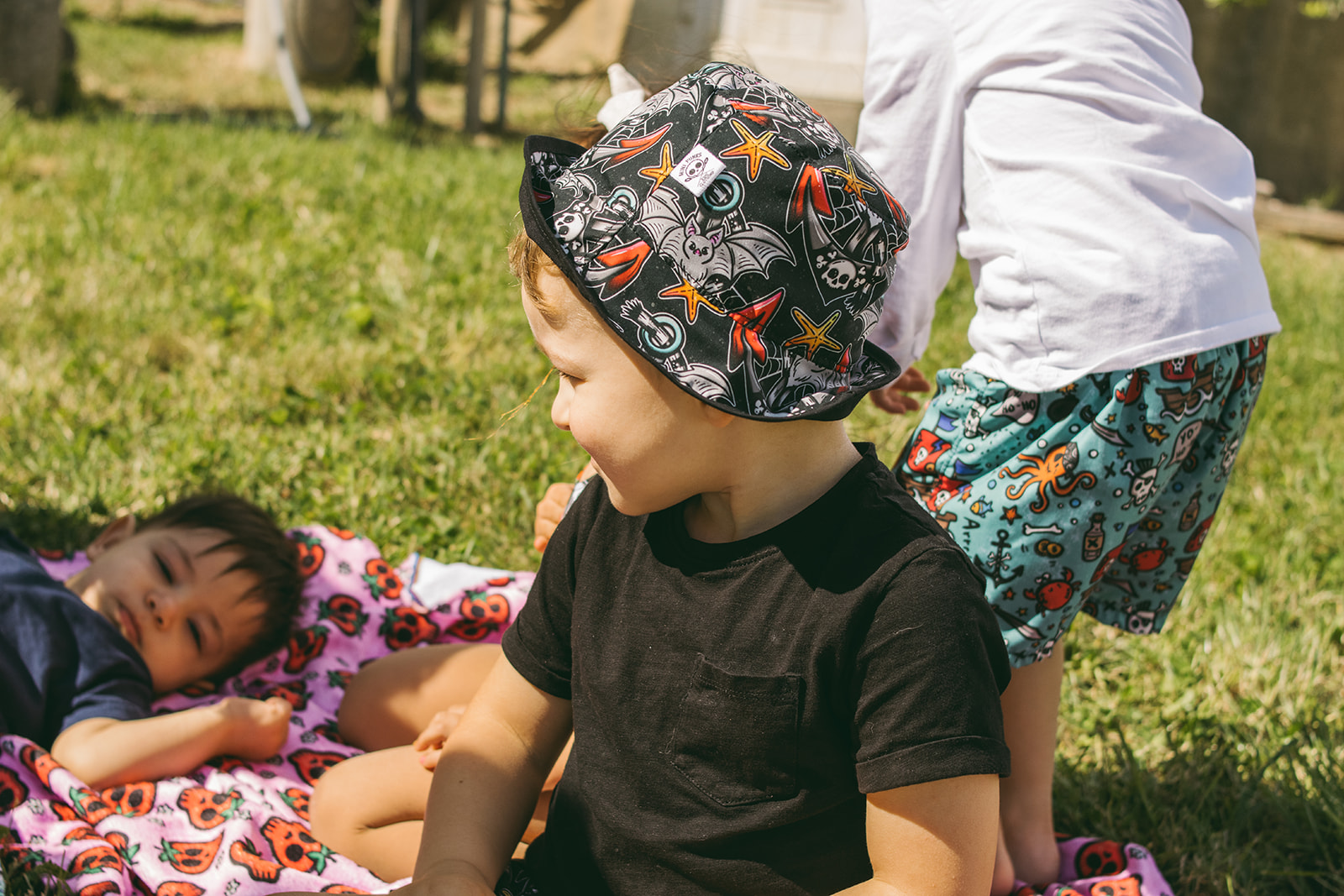 Criança com chapéu estampado e camiseta preta a brincar numa relva com outras crianças.