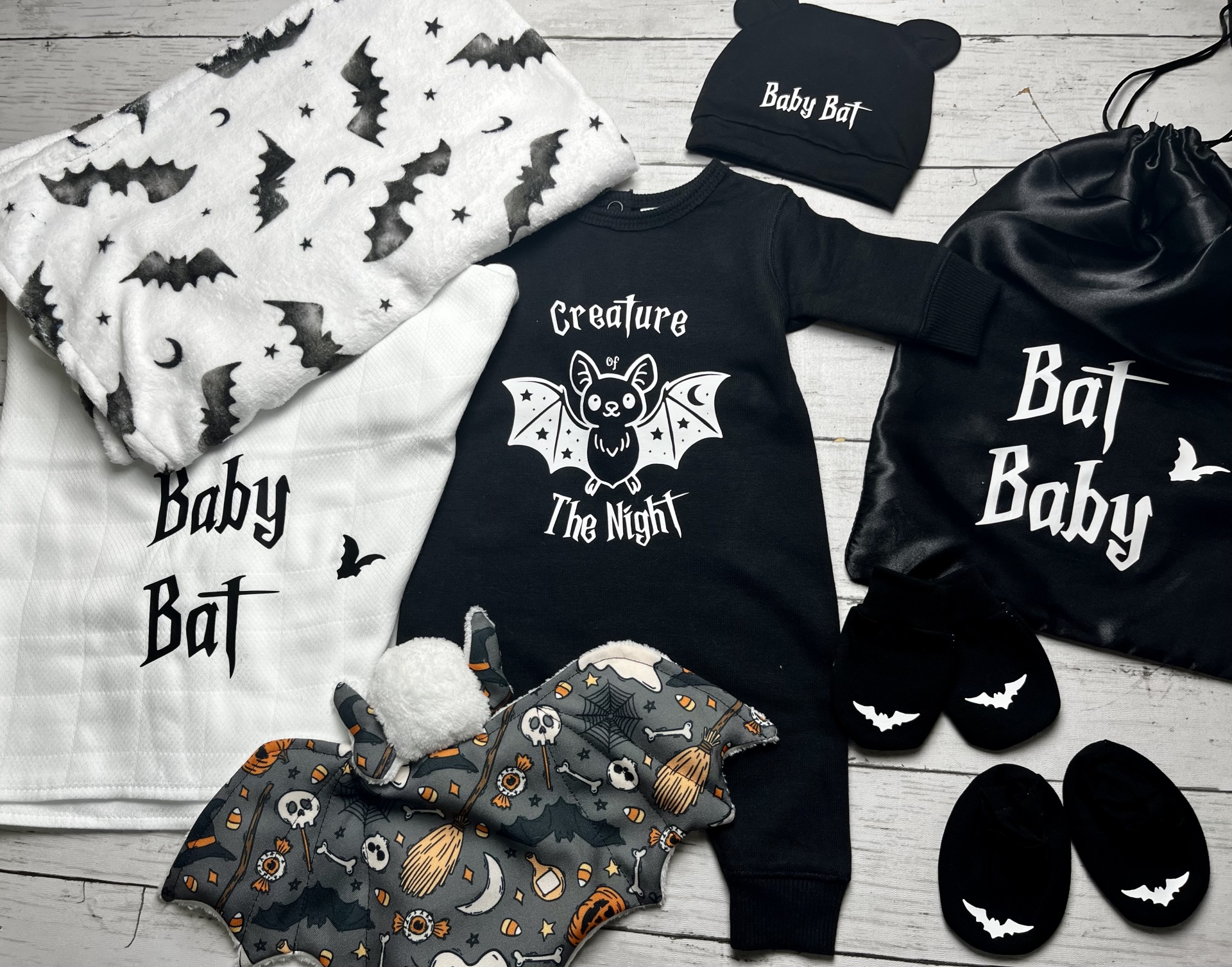 Roupas e acessórios para bebé com tema de morcego em cores preto, branco e cinzento, incluindo pijama, touca, sapatinhos, saco, manta e toalha