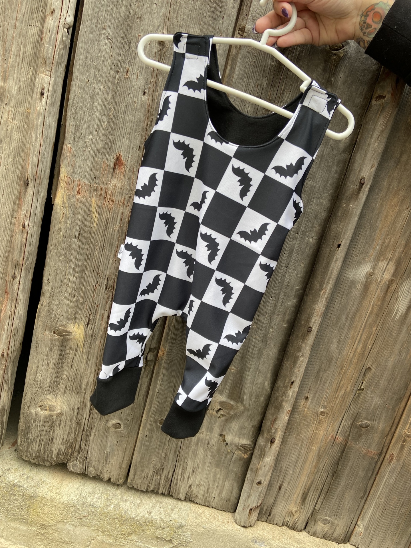 Macacão preto e branco com padrão de morcegos, pendurado em cabide branco, em porta de madeira.