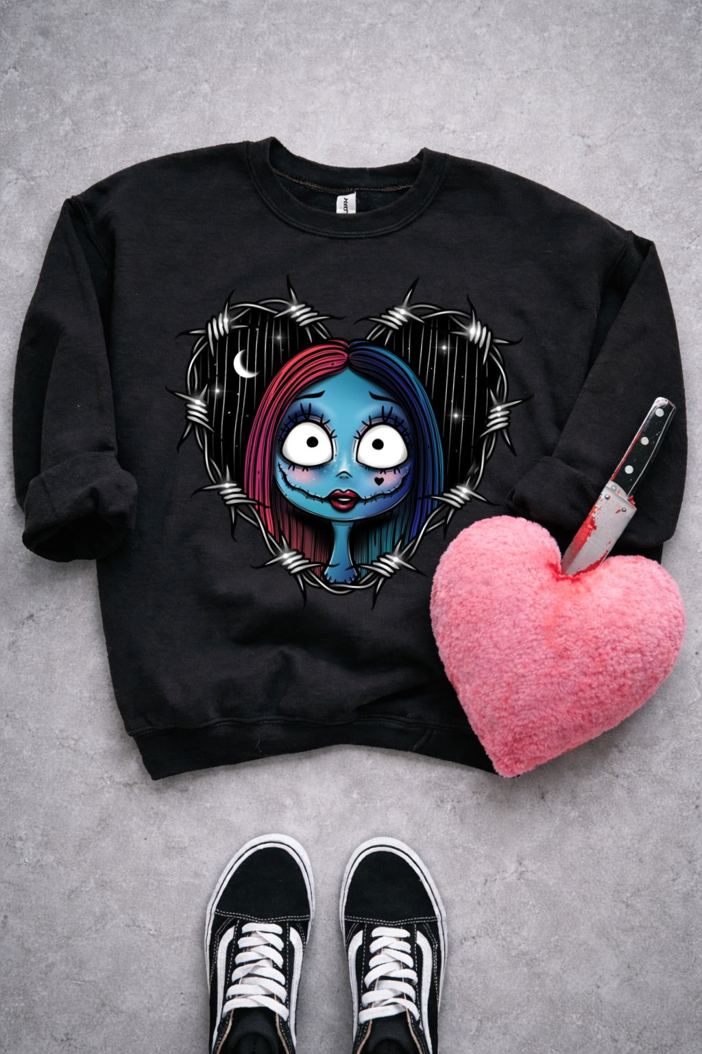 Sweatshirt preta com estampa colorida de rosto feminino em coração de arame farpado, coração de peluche rosa com faca e ténis pretos no chão.