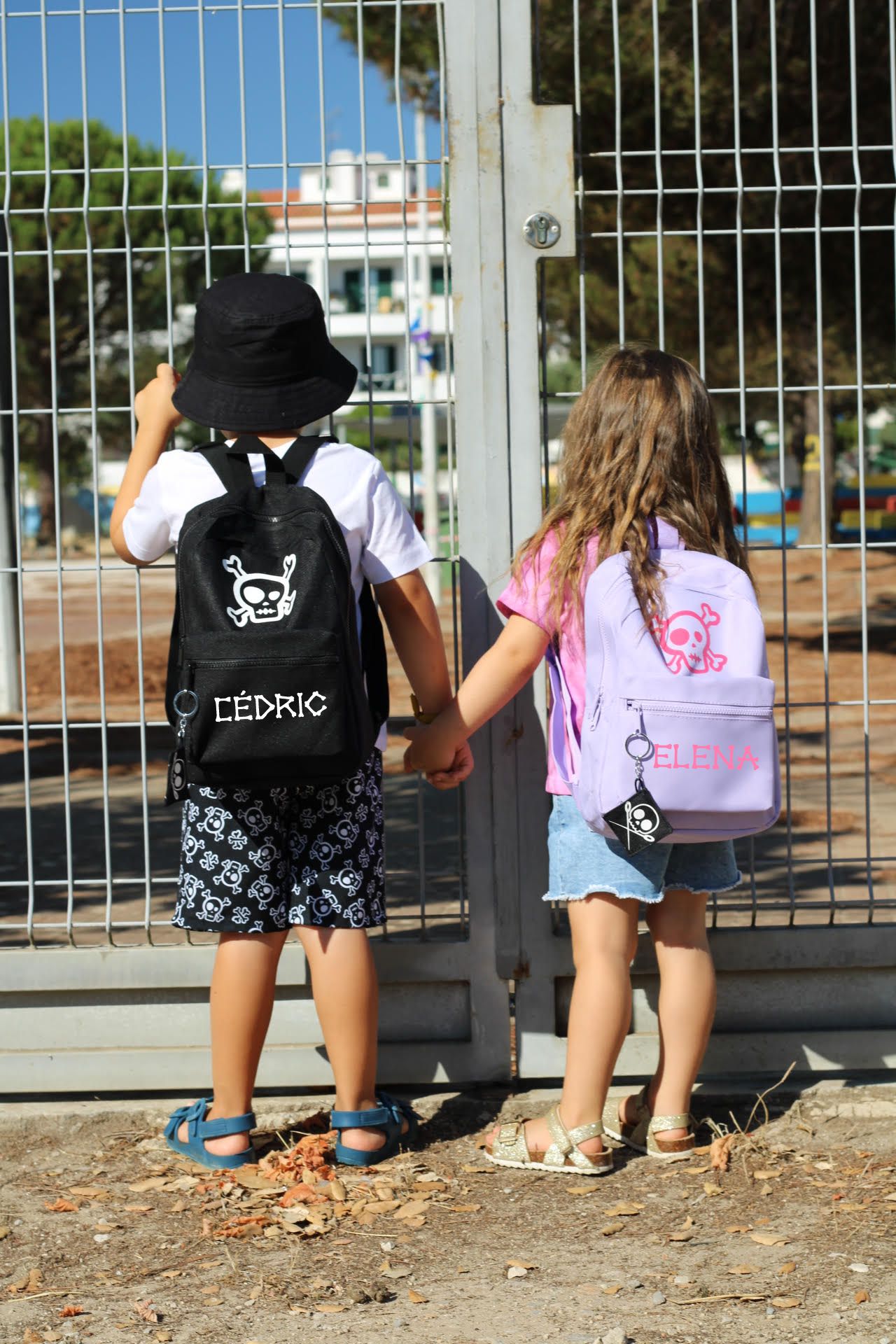 Crianças de costas com mochilas personalizadas, diante de grade metálica.