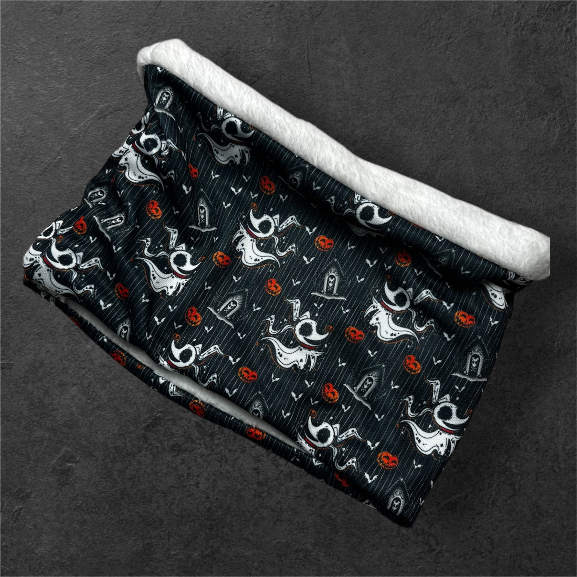 Cachecol tubular preto com padrão de fantasmas, abóboras e lápides, interior branco felpudo.