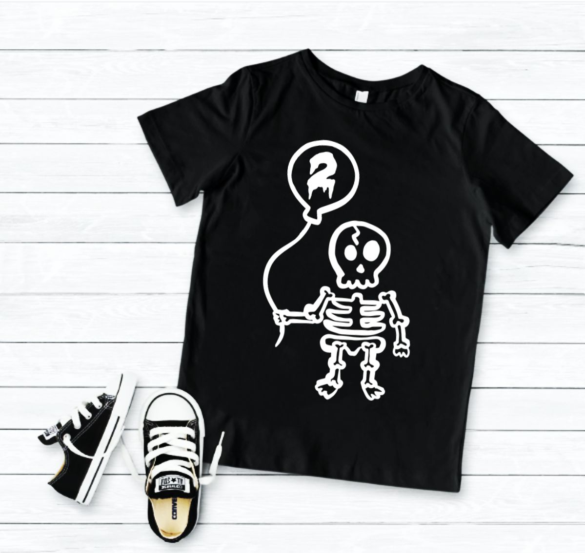 T-shirt preta com estampa de esqueleto e balão, ténis infantis pretos e brancos sobre chão de madeira clara