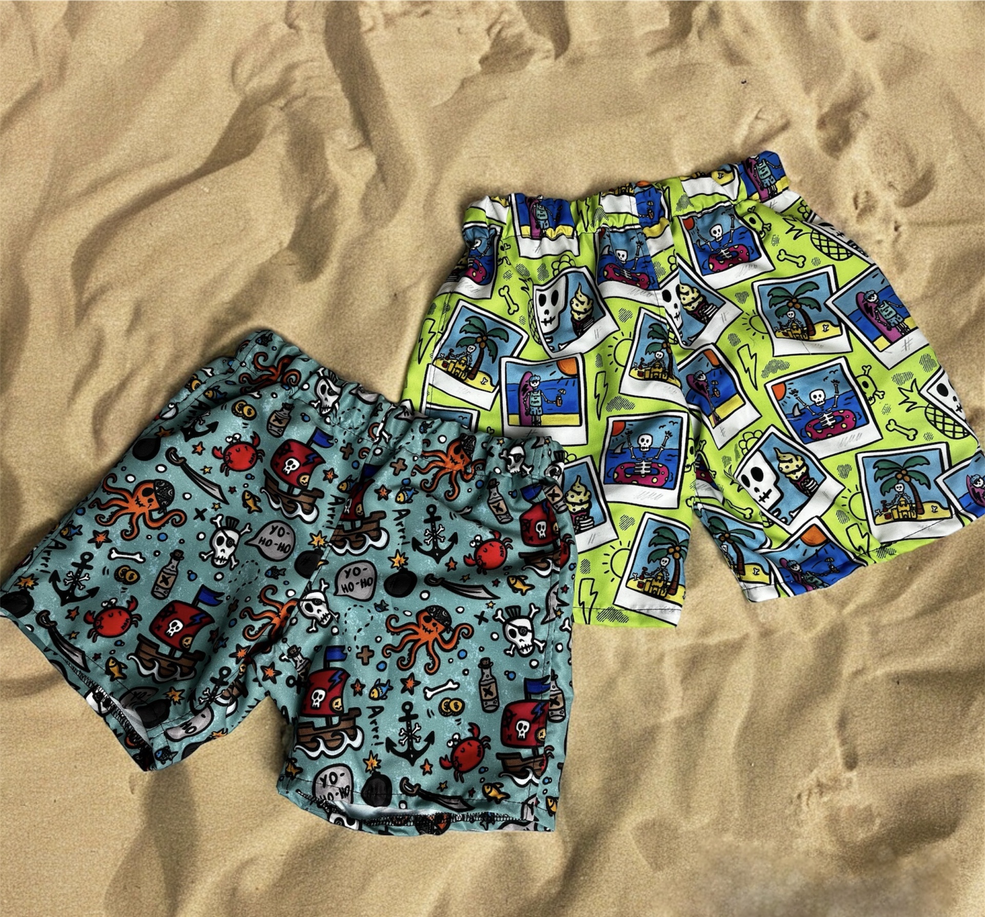 Dois calções de banho infantis com padrões coloridos sobre areia.