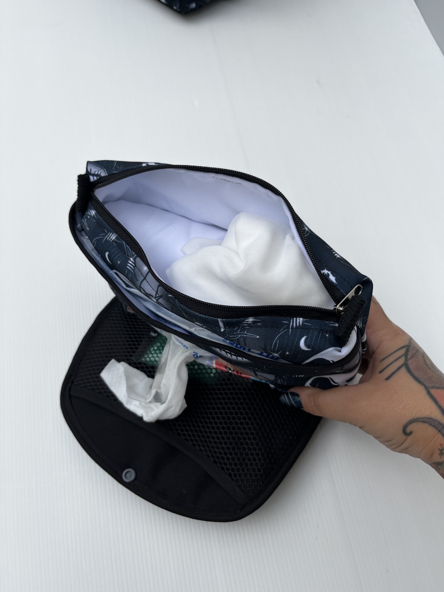 Bolsa organizadora azul escura aberta com tecidos brancos, mão tatuada segurando, sobre superfície branca e bolsa preta com lenços