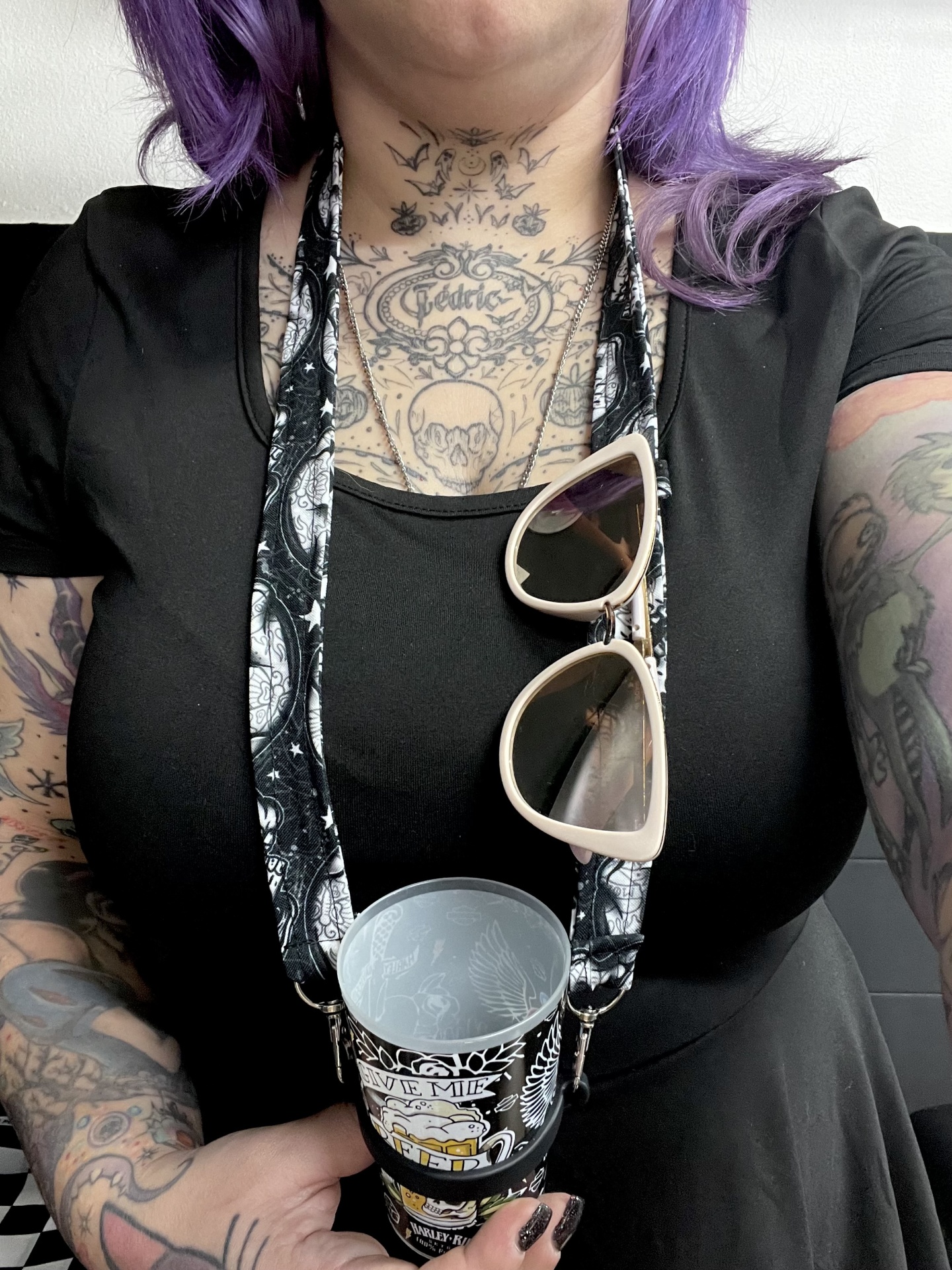 Mulher com tatuagens, vestido preto, segura copo com alça preta e óculos de sol bege