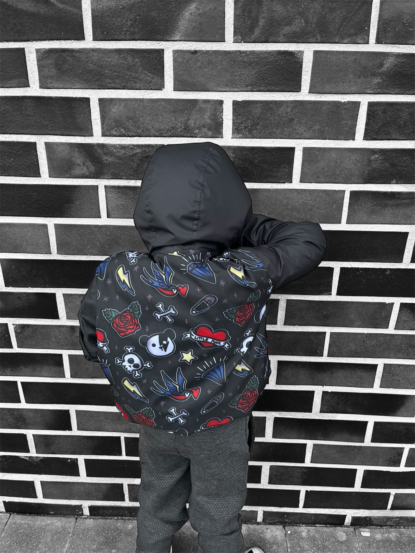Criança de costas com casaco preto estampado encostada a parede de tijolos pretos e cinzentos