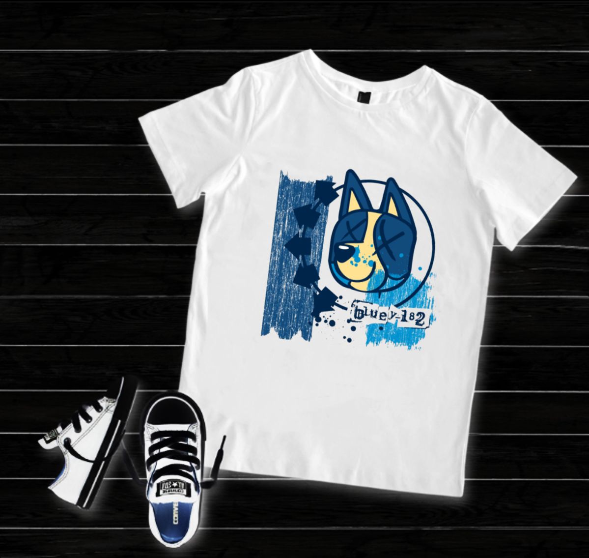 Camiseta branca com estampa de cão azul e amarelo, sapatos desportivos brancos e pretos sobre chão de madeira escura