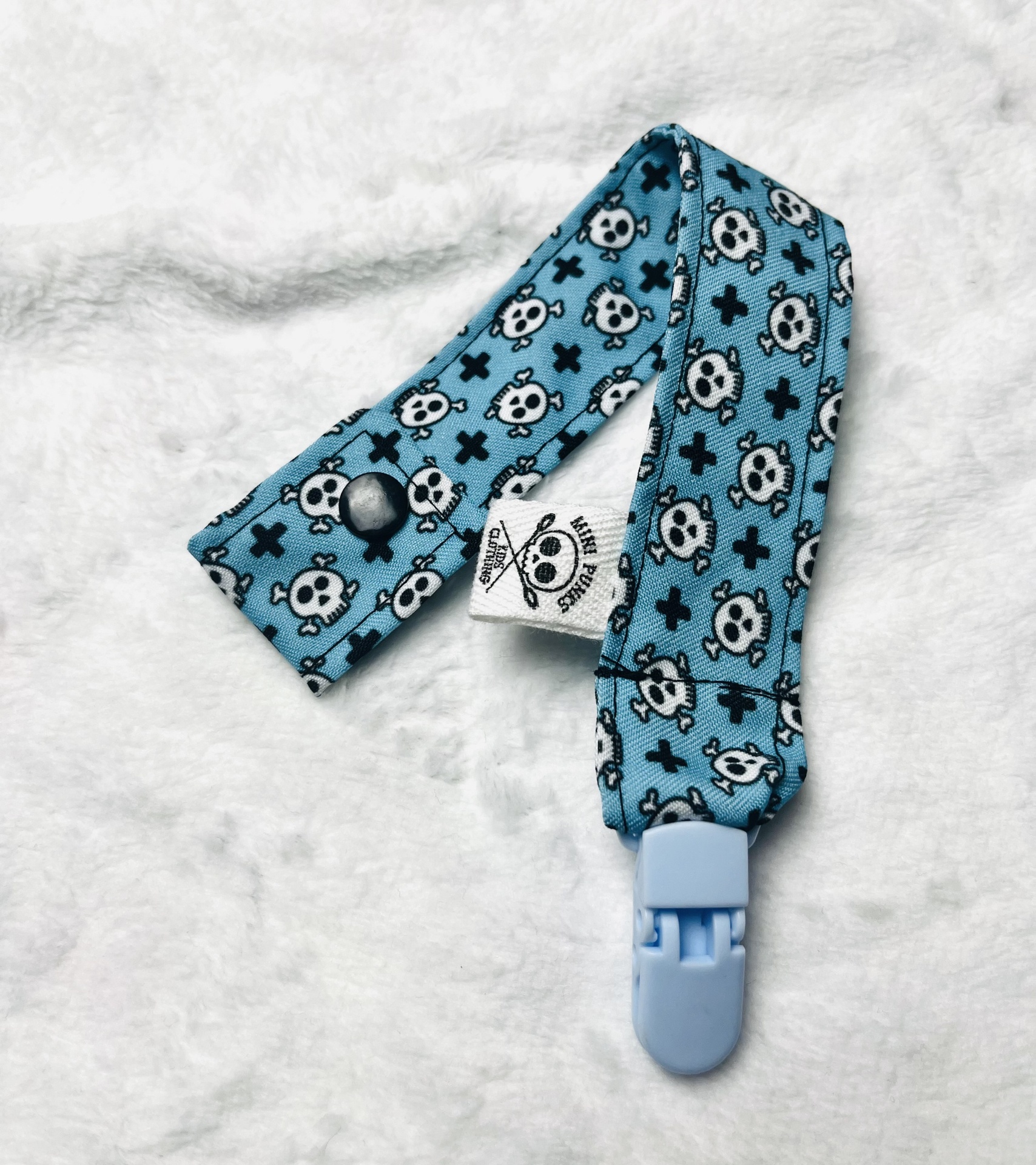Prendedor de chupeta azul com caveiras brancas e cruzes pretas.