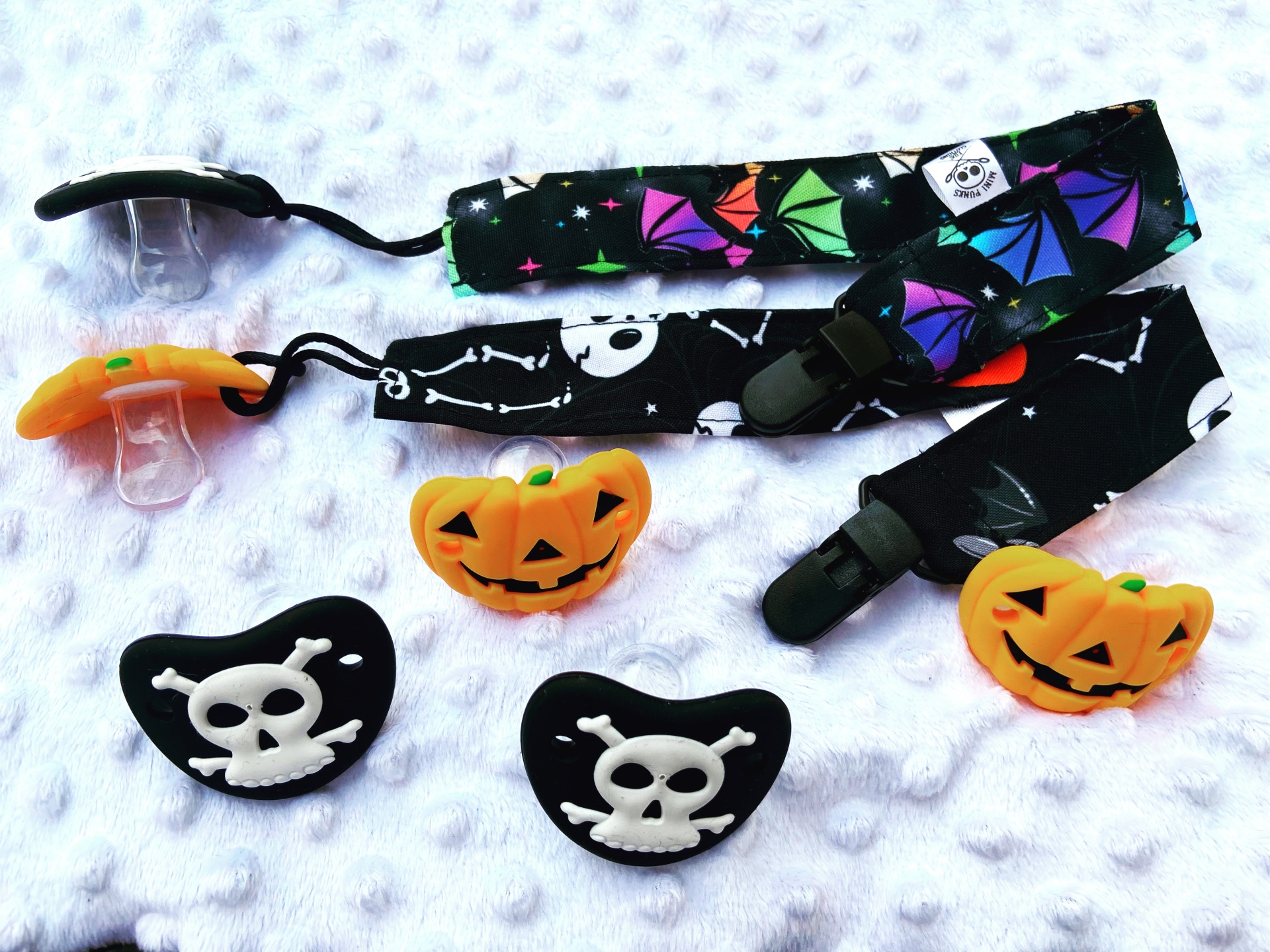 Chupetas com clips estampados e protetores em forma de abóboras e caveiras sobre fundo branco