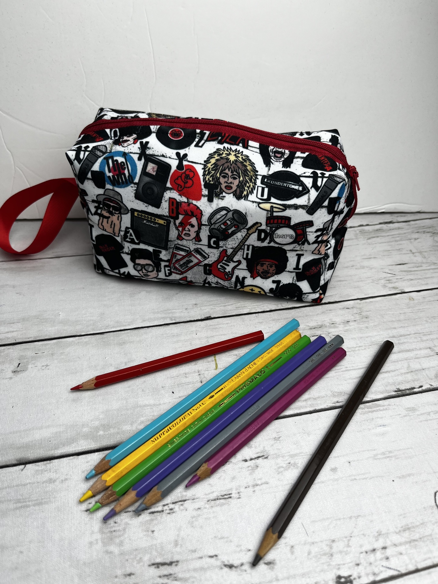 Necessaire branca com padrão colorido de desenhos musicais e fecho vermelho, junto a lápis de cor sobre mesa branca