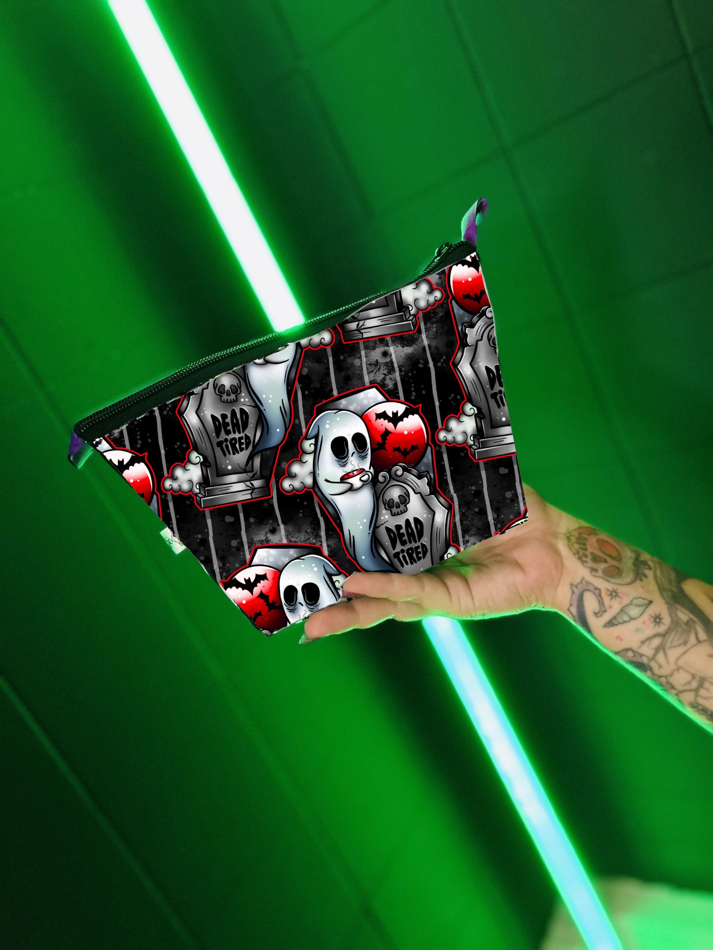 Necessaire preta com padrão de fantasmas e lápides, texto 'DEAD TIRED', segurada por mão tatuada