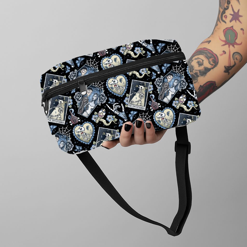 Bolsa tiracolo preta com padrão de caveiras e cartas, fecho e alça preta, segurada por mão com unhas negras e braço tatuado.