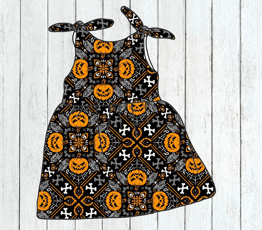 Vestido infantil preto com padrão de abóboras, teias de aranha e cruzes, alças em laço, sobre madeira clara.