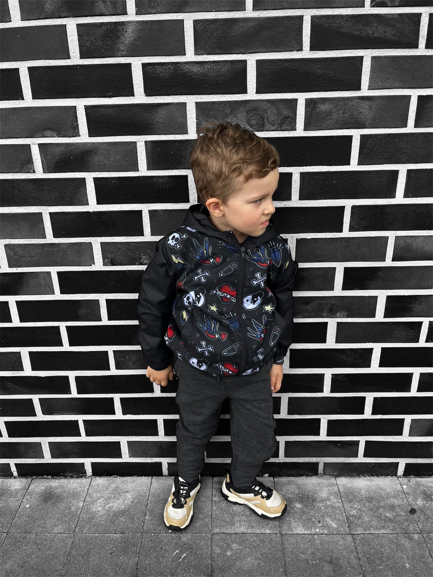 Criança com casaco preto estampado e calças cinza em frente a parede de tijolos pretos
