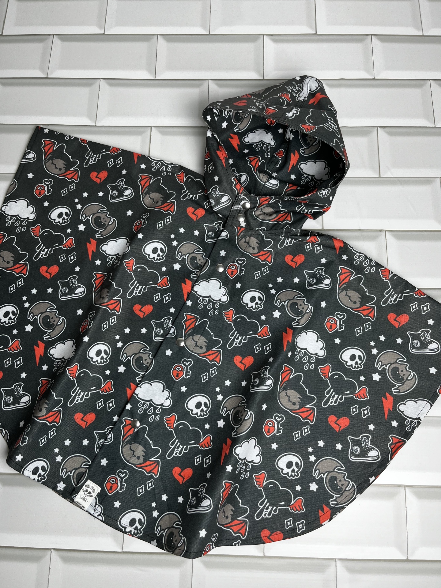 Poncho preto com capuz com padrão de caveiras, corações partidos, morcegos e nuvens, tecido impermeável sobre azulejos brancos.