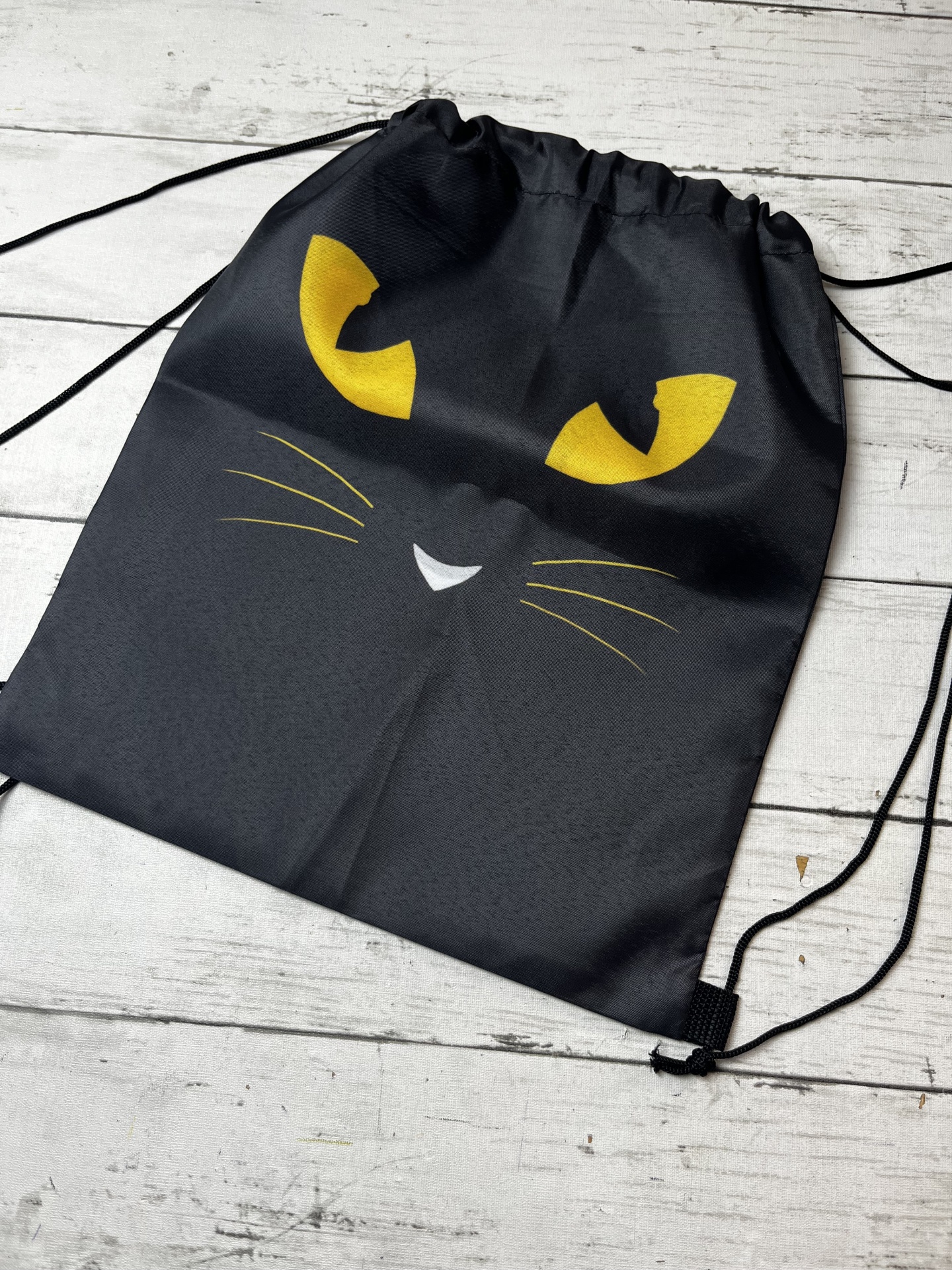 Mochila preta com rosto de gato em tecido, cordões pretos, sobre madeira clara.
