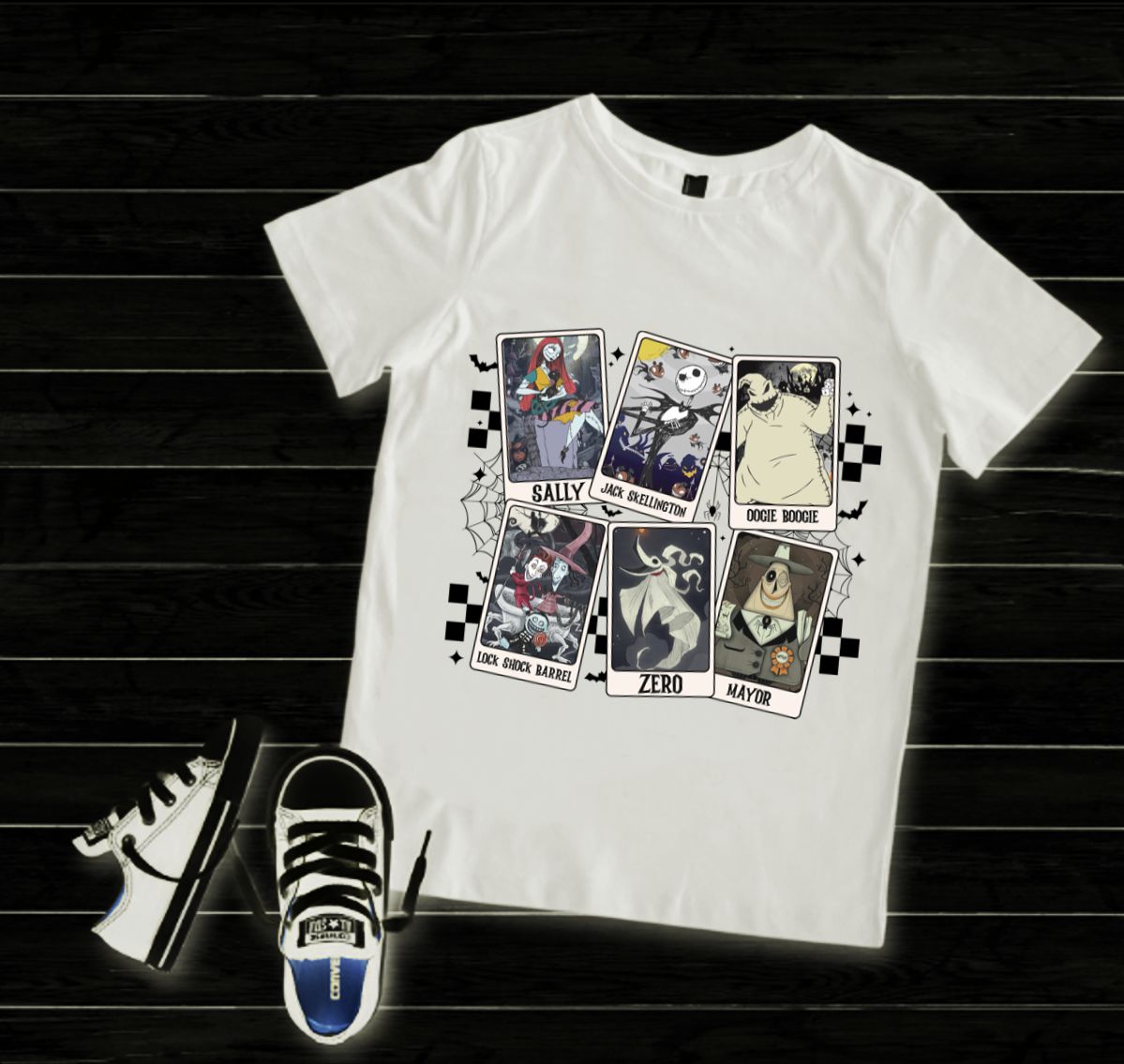 T-shirt branca com estampas coloridas de personagens do filme O Estranho Mundo de Jack junto a sapatilhas pretas e brancas sobre piso de madeira preto.