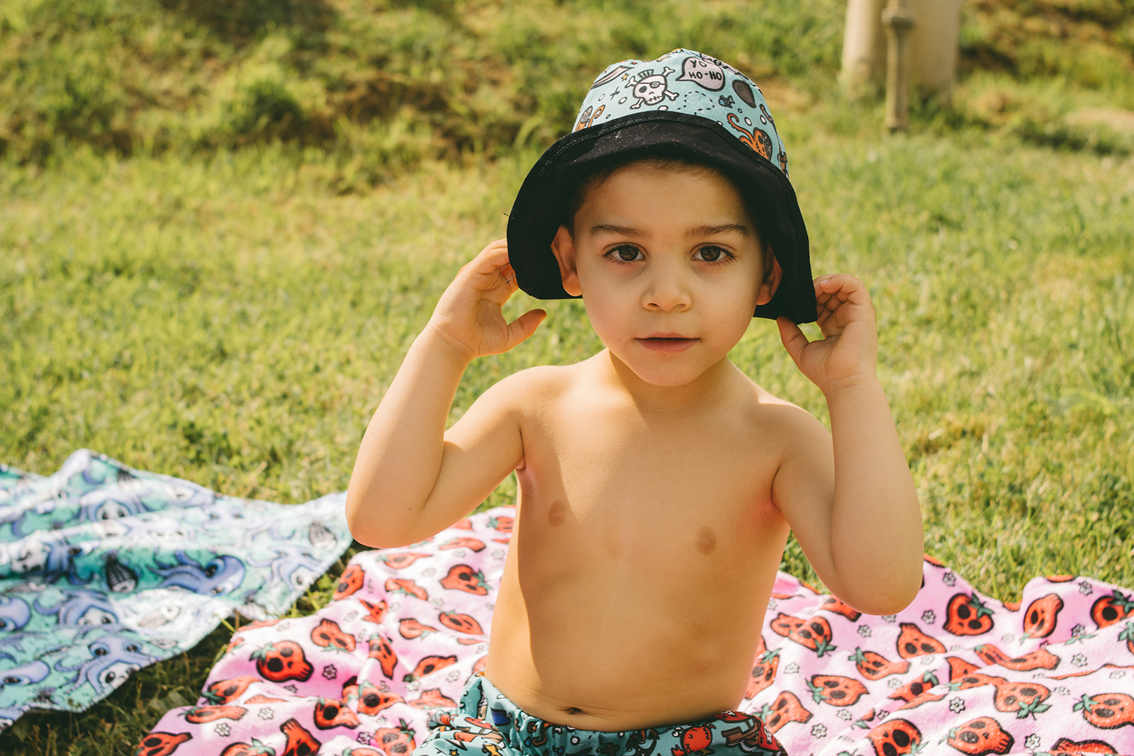 Criança com chapéu azul sentado em mantas coloridas com desenhos de caveiras em relvado