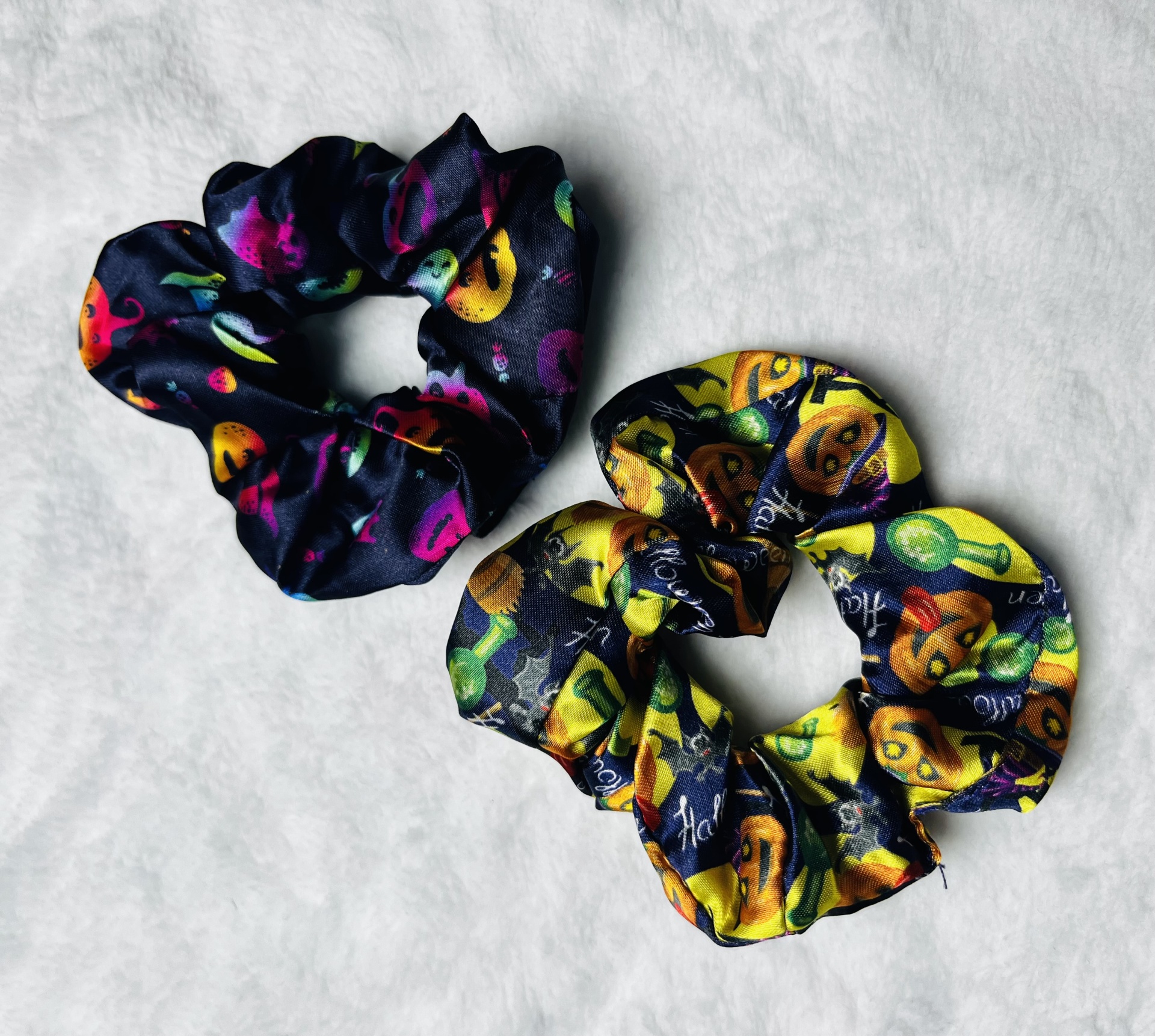 Dois scrunchies com padrão Halloween apresentando abóboras e morcegos, um com fundo preto e outro amarelo, sobre superfície branca.