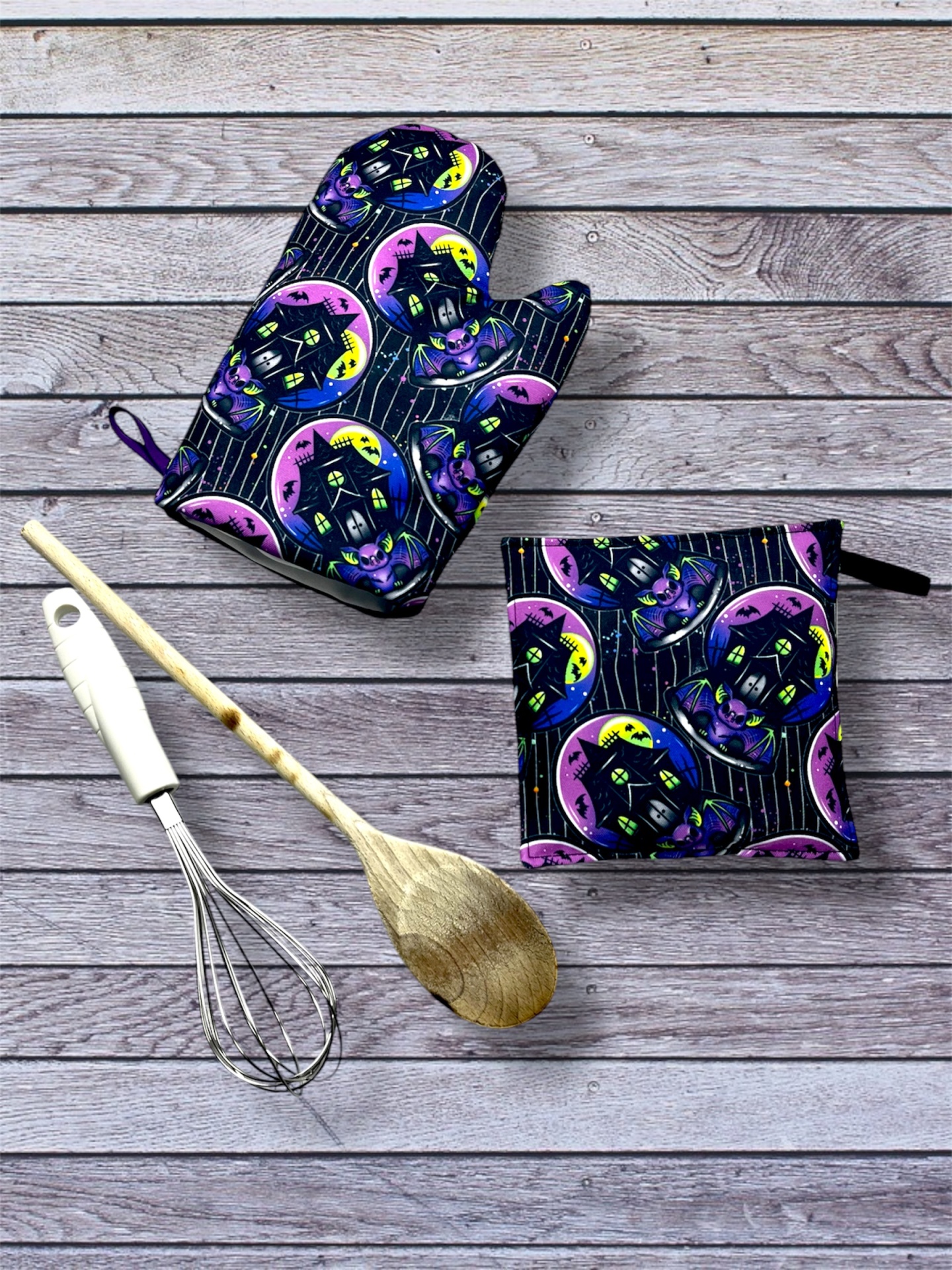 Luvas e pega-luvas de cozinha com padrão de casas amarelas e roxas sobre fundo listrado preto ao lado de utensílios de cozinha sobre madeira cinza