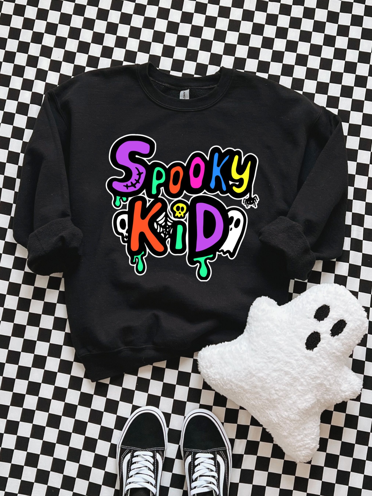 Sweatshirt preta com estampa Spooky Kid colorida sobre piso xadrez, peluche de fantasma branco e ténis pretos.