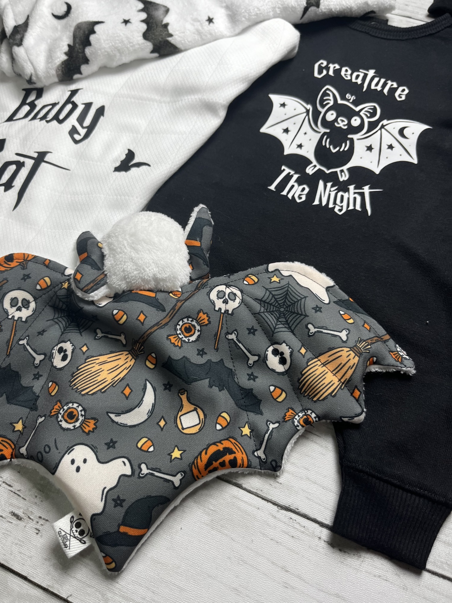 Conjunto para bebé com padrão de morcegos e texto em algodão branco, body preto com morcego e texto, e peluche Halloween colorido