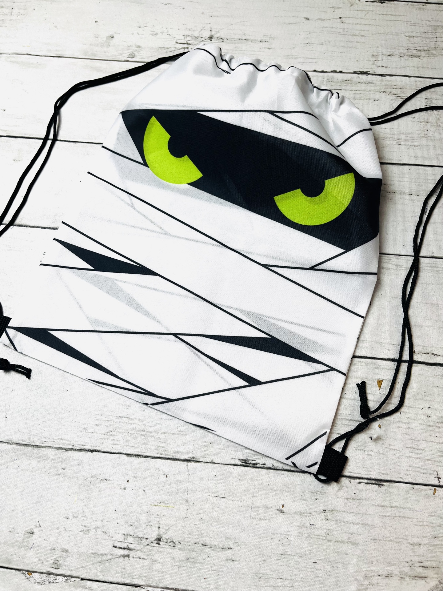 Mochila de cordões branca com olhos amarelos e pretos desenhados, sobre fundo de madeira clara