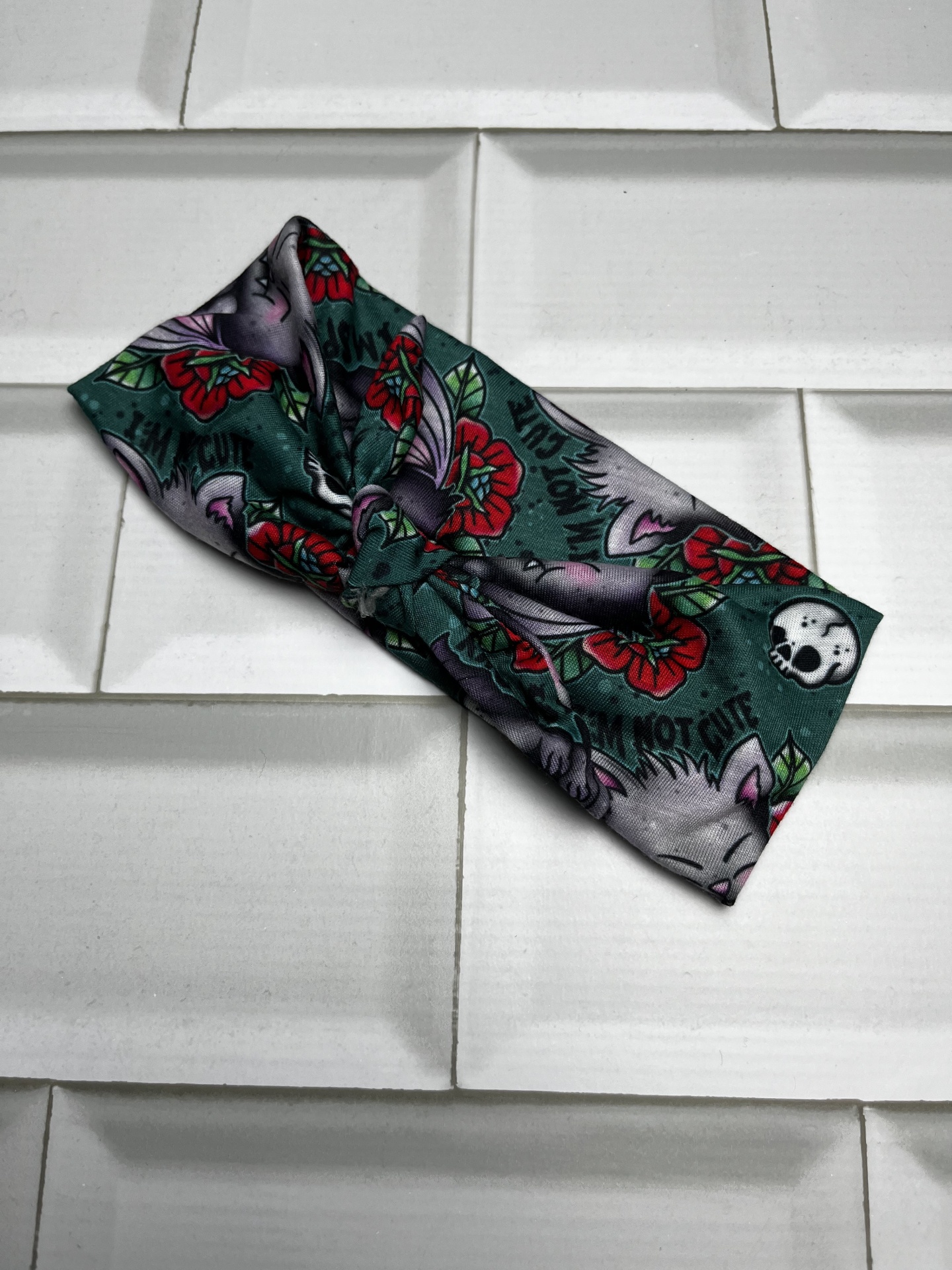 Faixa de cabelo com padrão de gatos, flores vermelhas, folhas verdes, texto 'I AM NOT CUTE' e caveira em azulejos brancos.