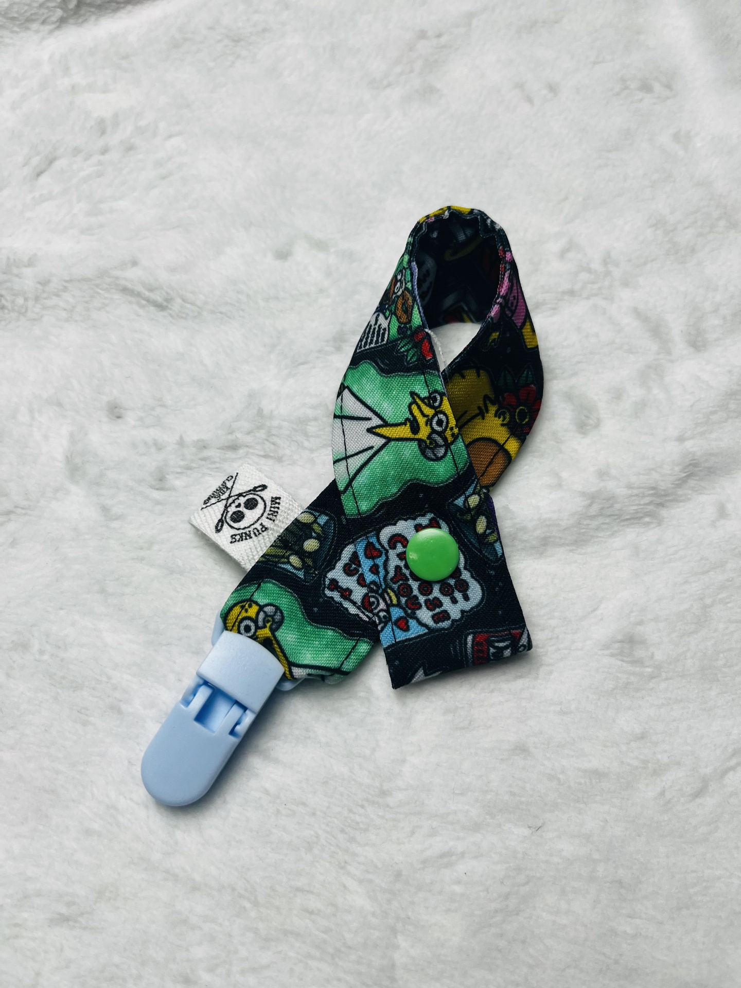 Prendedor colorido com padrão cartoon e clipe azul sobre fundo branco felpudo.