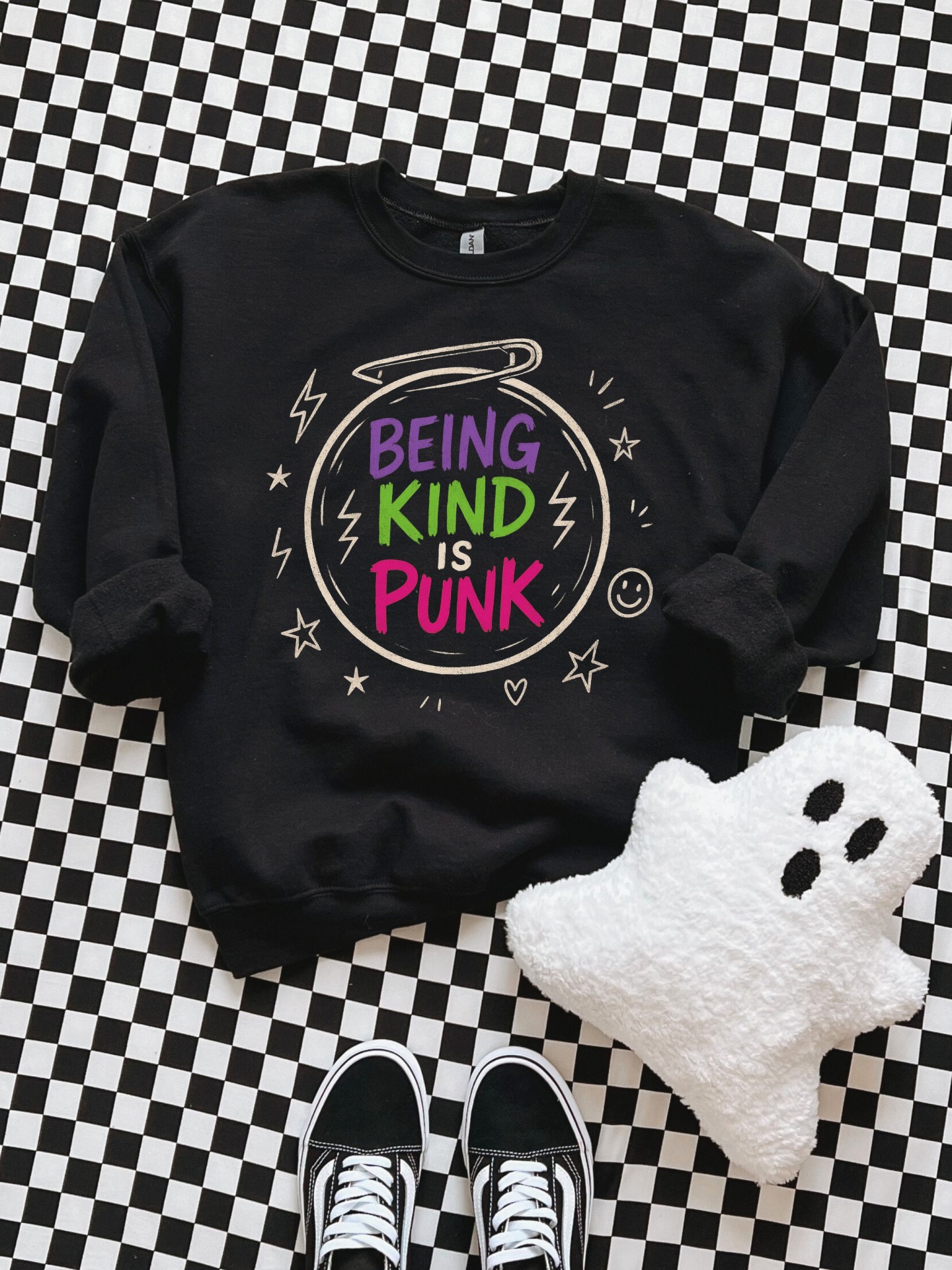 Sweatshirt preta com frase colorida BEING KIND IS PUNK sobre fundo xadrez, perto de almofada fantasma e sapatos pretos com atacadores brancos.