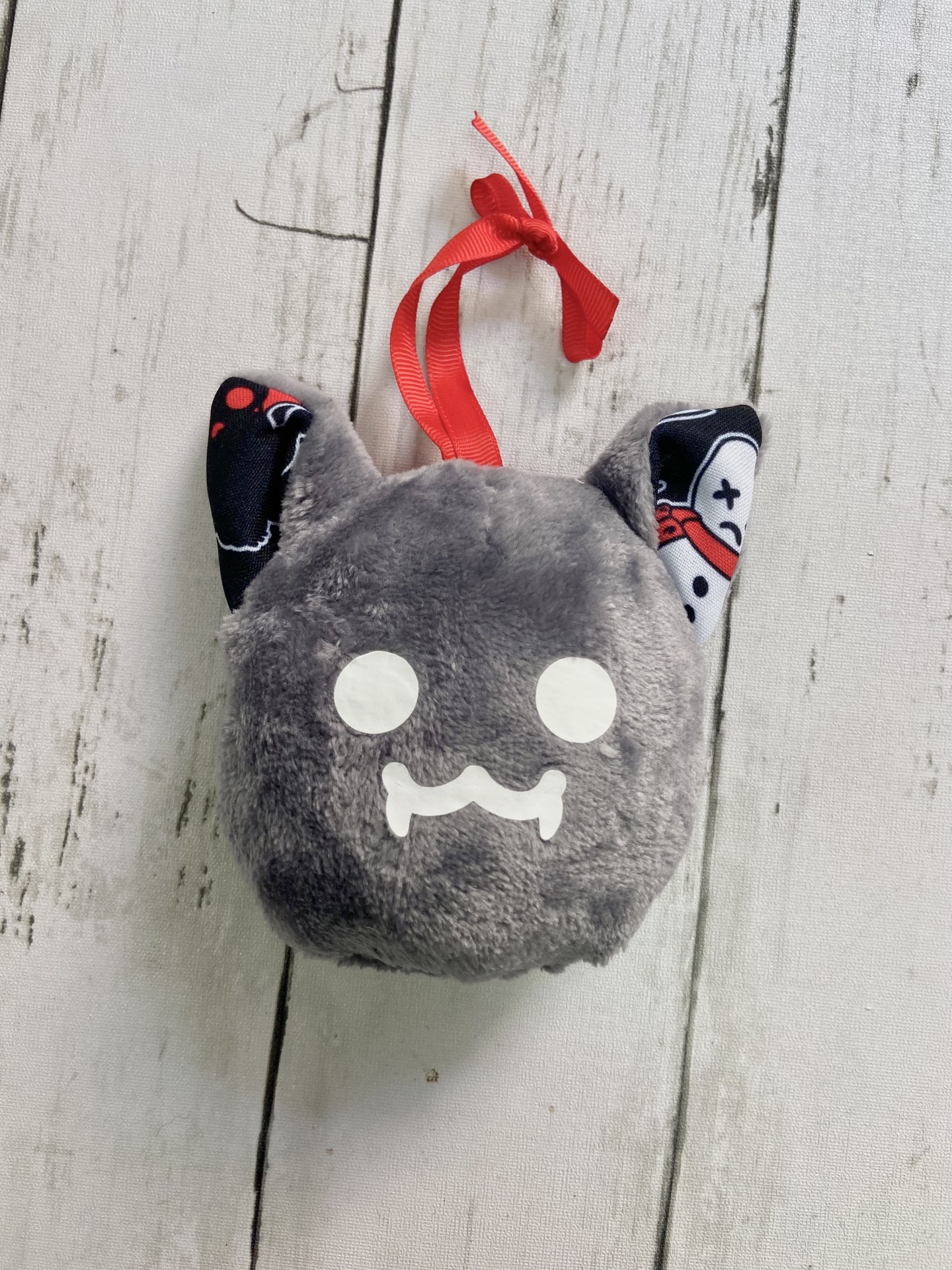 Pelúcia cinzenta com rosto branco e orelhas pretas decoradas pendurada numa fita vermelha sobre fundo de madeira clara