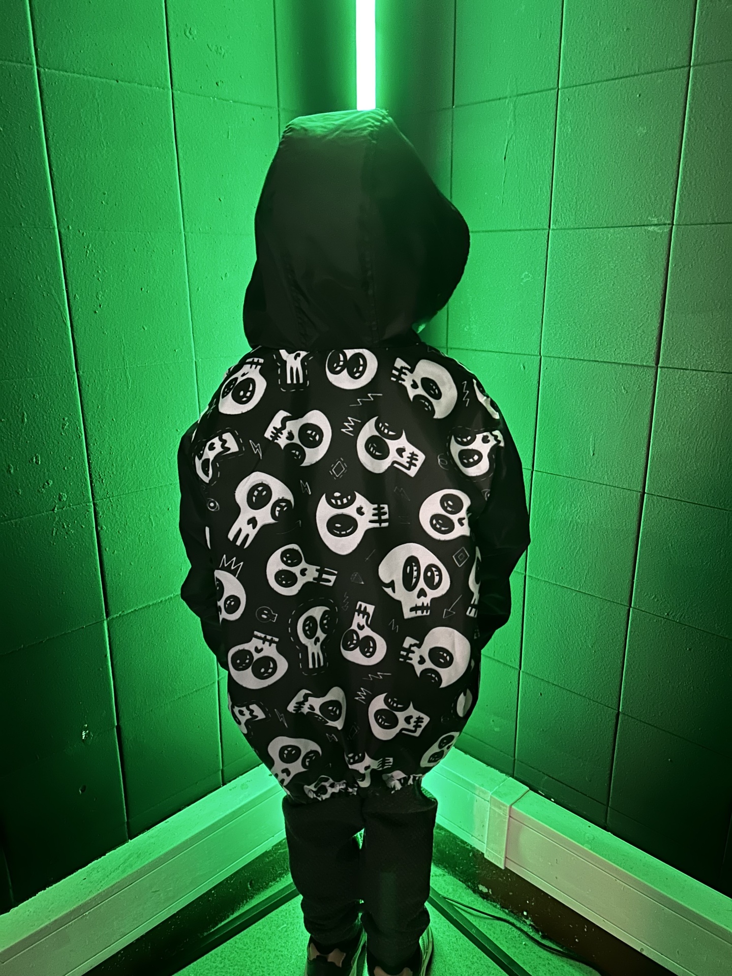 Criança de costas a usar macacão preto com padrão de caveiras brancas, em ambiente com luz verde