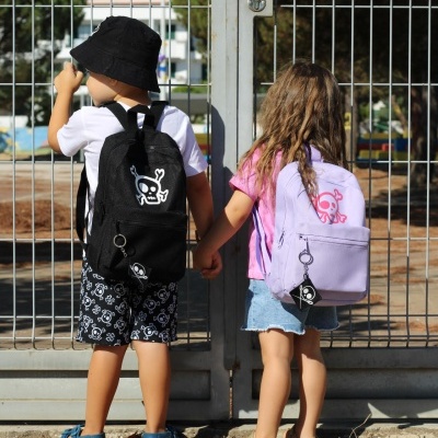 Duas crianças de costas com mochilas e roupas casuais, segurando as mãos em frente a uma grade metálica branca.