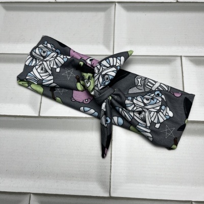 Faixa de cabelo preta com padrão de múmias e estrelas coloridas sobre azulejos brancos