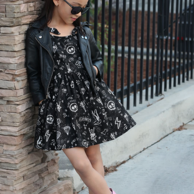 Criança com vestido preto estampado, casaco de couro preto, botas rosas, laço de cabelo rosa e óculos escuros encostada numa parede de pedra