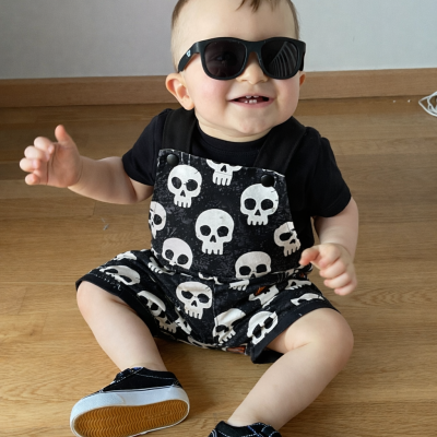 Bebé com macacão de caveiras, óculos escuros e sapatilhas sentado no chão de madeira