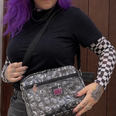 Mala tiracolo preta e cinzenta com padrão de caveiras e emblema rosa com texto, usada por pessoa com cabelo roxo